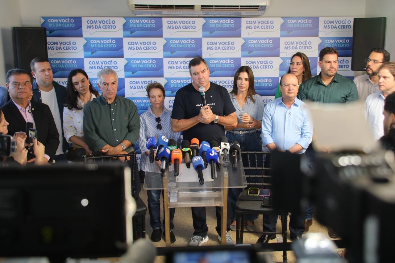 Um homem de camiseta preta fala ao microfone em uma coletiva de imprensa, rodeado por outras pessoas e microfones de diversas mídias. Ao fundo, um painel azul e branco com a frase Com você o MS dá certo