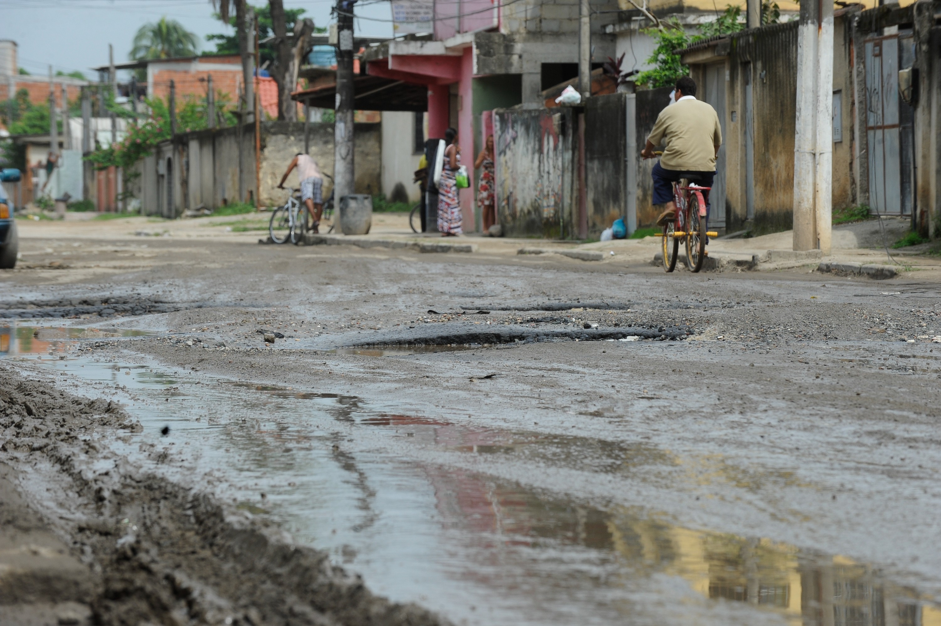 Rua de terra batida com muitas poças e buracos, refletindo o céu nublado. Um homem de camisa bege e calça escura pedala uma bicicleta vermelha à direita, enquanto outro homem de camiseta branca e bermuda empurra uma bicicleta à esquerda. Ao fundo, casas simples e muros de concreto compõem o cenário urbano