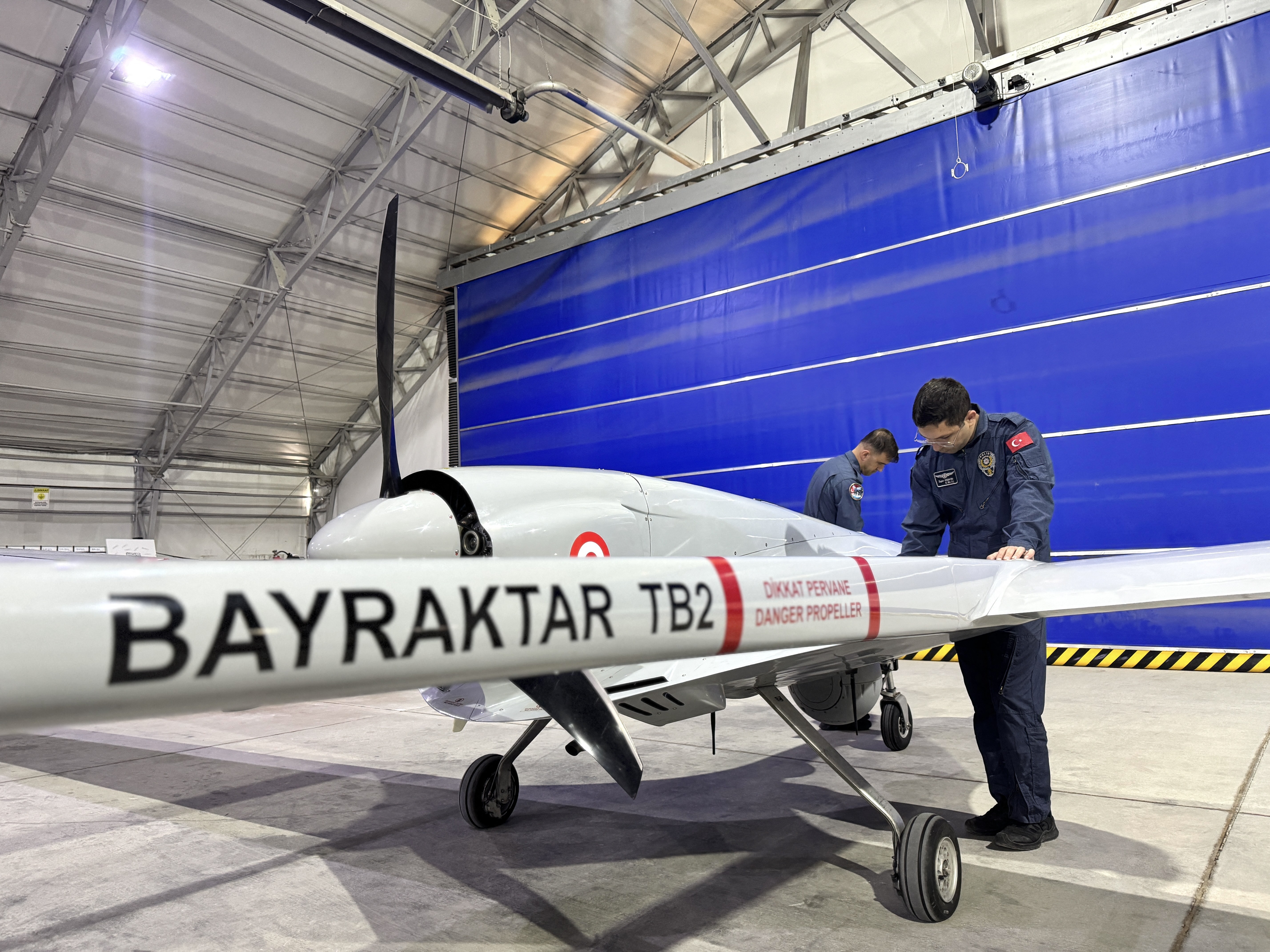 Dois homens de uniforme azul inspecionam um drone militar branco Bayraktar TB2 com hélice traseira, dentro de um hangar com teto metálico e parede azul
