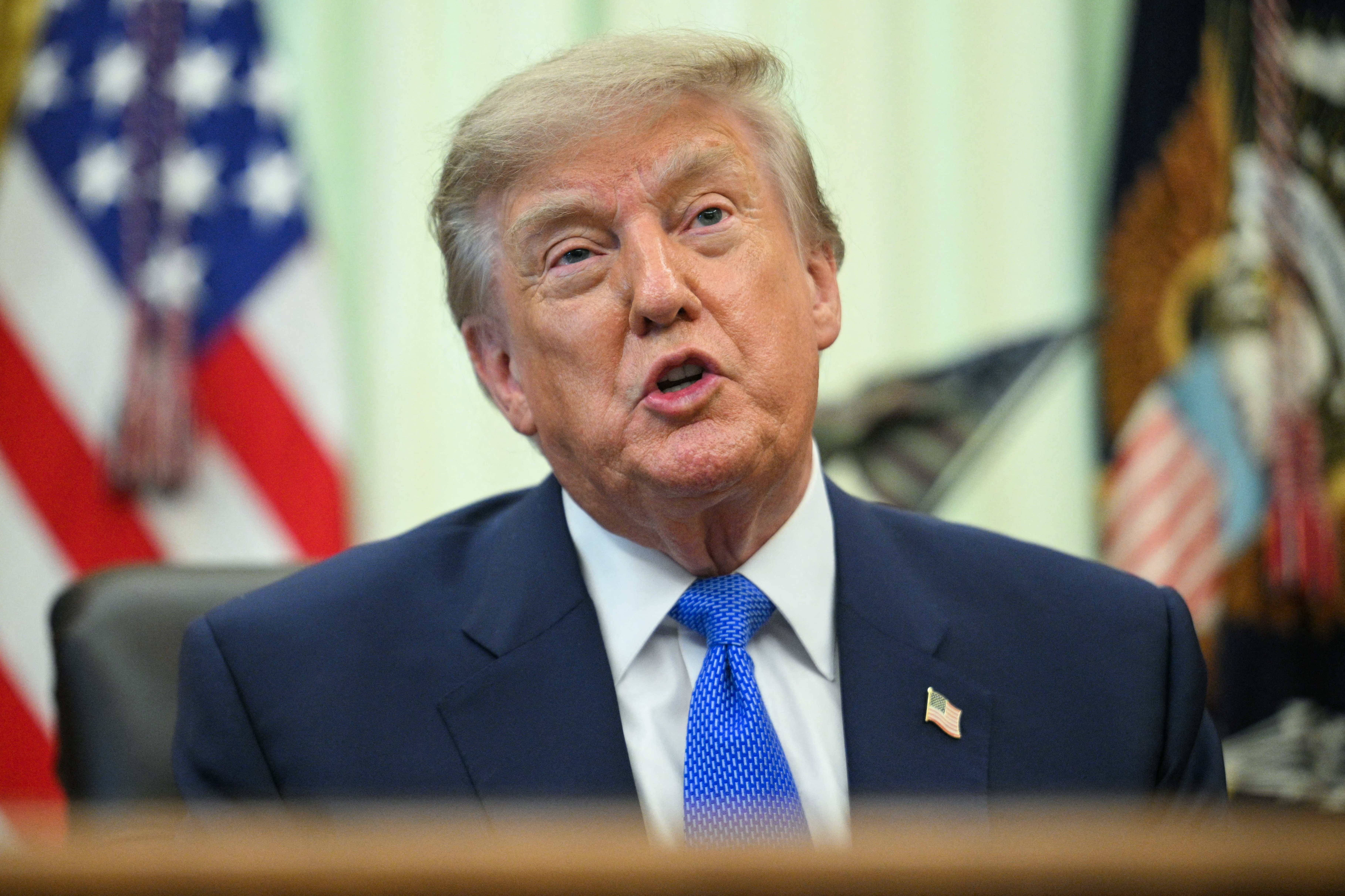 Donald Trump, homem branco de cabelos loiros, vestindo terno azul-marinho, camisa branca e gravata azul, fala com a boca aberta. Ao fundo, desfocadas, bandeiras dos Estados Unidos