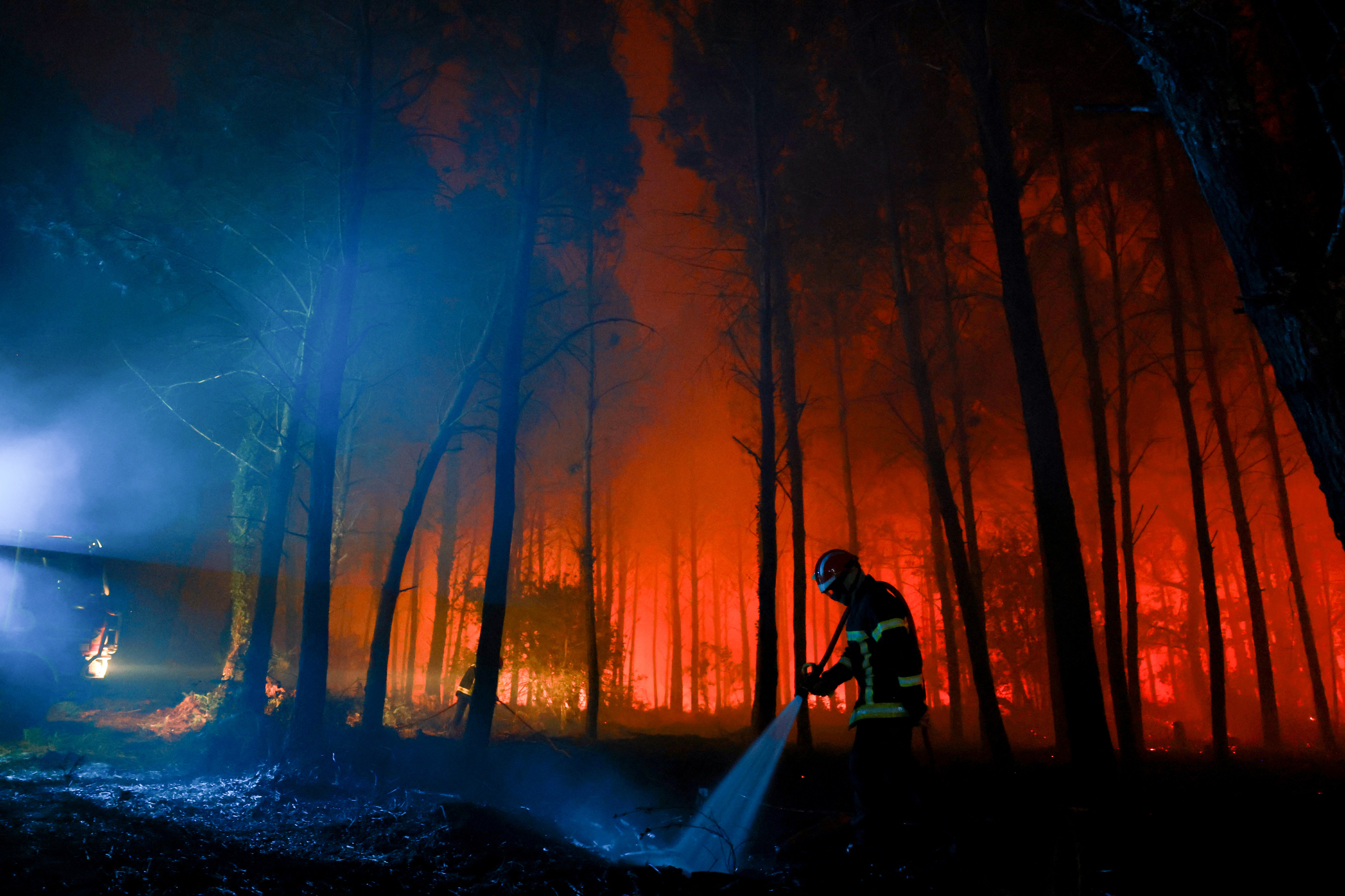 Bombeiro com capacete e uniforme escuro, com faixas refletivas, joga água no chão de uma floresta em chamas. As árvores altas e finas estão envoltas em fumaça vermelha e laranja do fogo, enquanto a área à esquerda é iluminada por uma luz azulada e fumaça branca, criando um contraste dramático