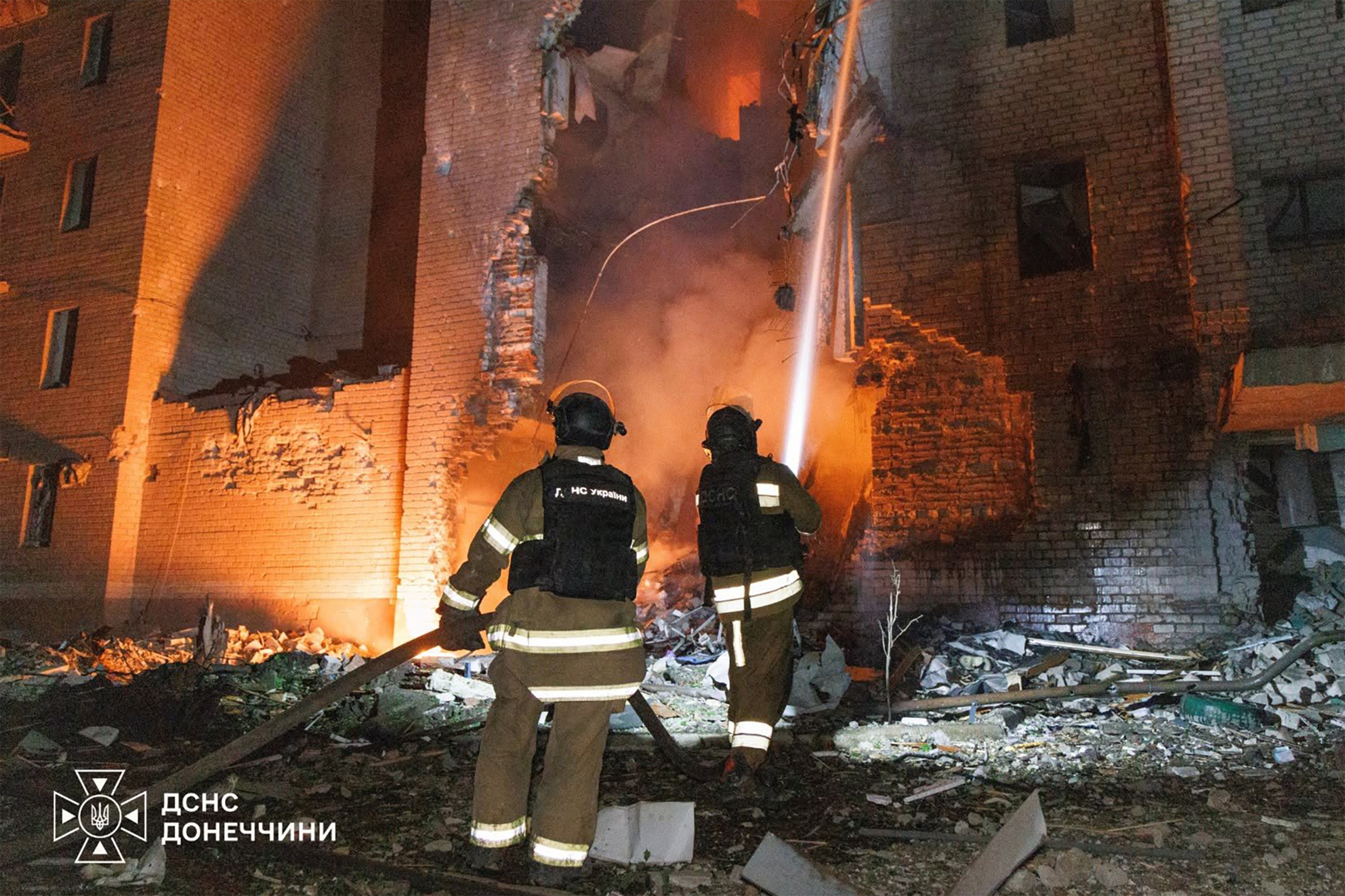 Dois bombeiros em uniforme de proteção, de costas, observam um prédio parcialmente destruído e em chamas, com fumaça e escombros no chão. Um jato de água é direcionado para o fogo. A cena noturna é iluminada pelo brilho alaranjado das chamas e por uma luz branca. No canto inferior esquerdo, o logo do Serviço de Emergência da Ucrânia para Donetsk