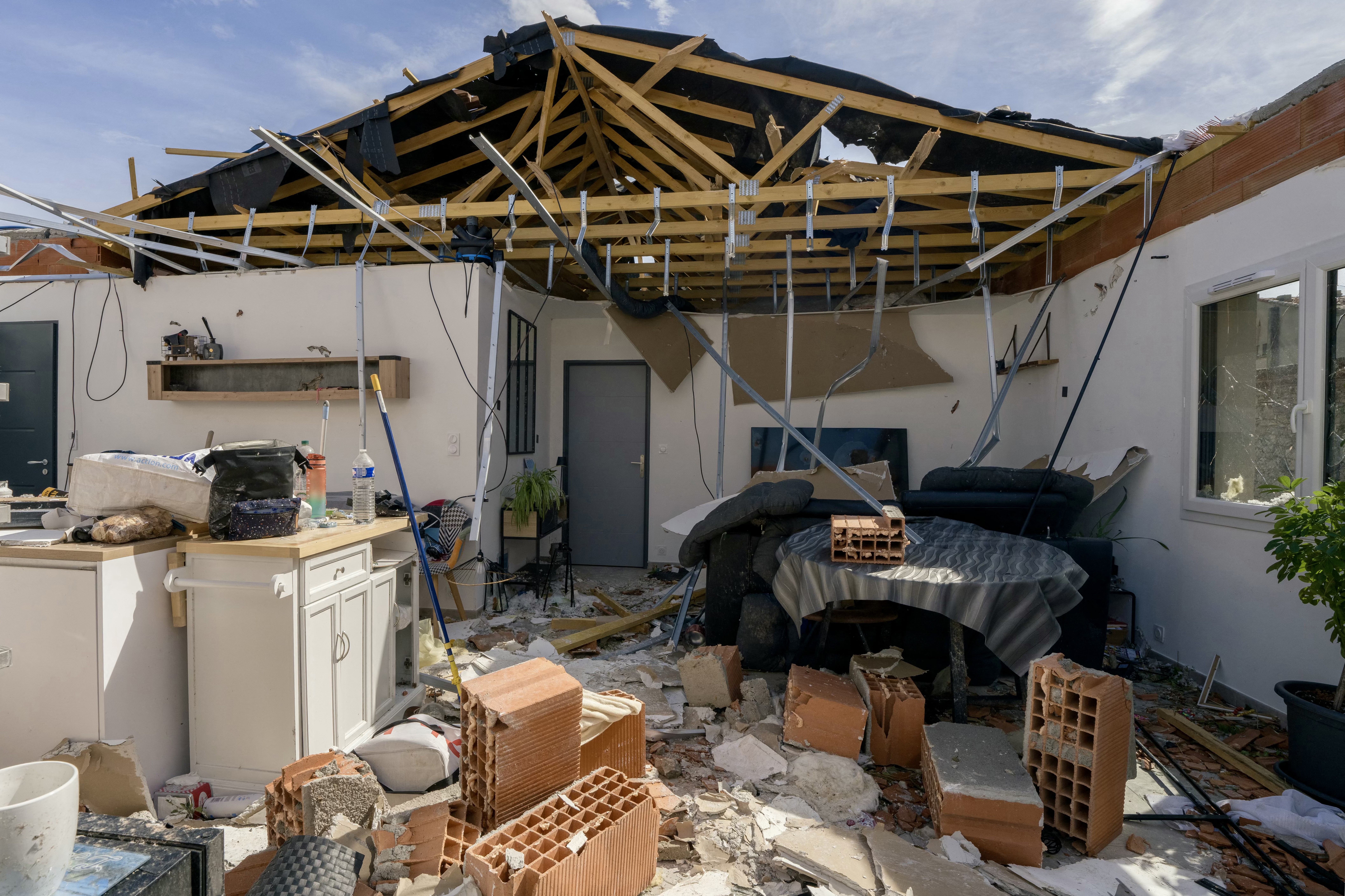 Interior de uma casa destru&iacute;da, com o telhado desabado expondo a estrutura de madeira e o c&eacute;u azul. Tijolos, escombros e m&oacute;veis revirados cobrem o ch&atilde;o, incluindo uma mesa coberta e um sof&aacute; escuro. Uma bancada de cozinha branca com objetos espalhados e uma porta cinza ao fundo completam a cena de devasta&ccedil;&atilde;o