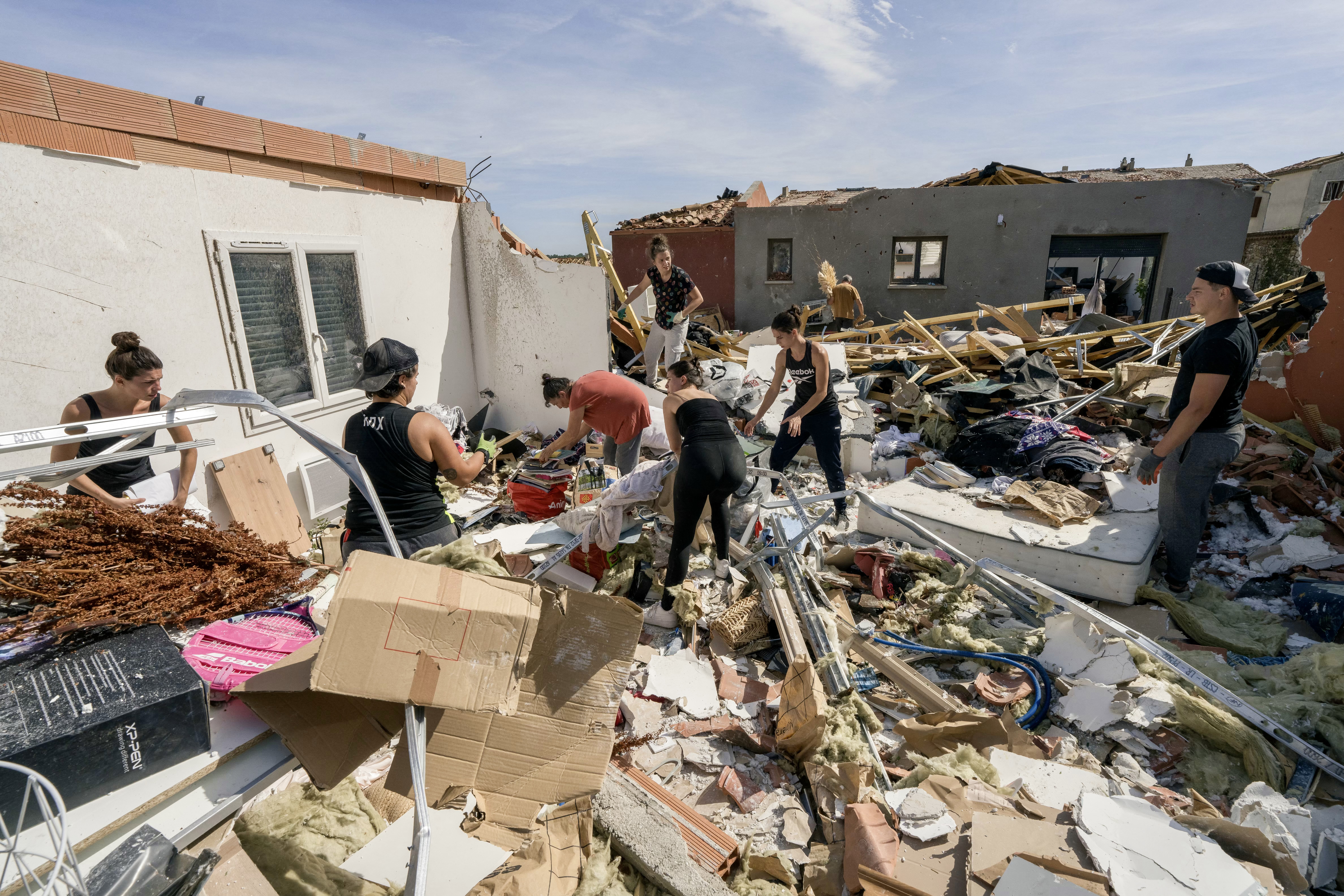Grupo de sete pessoas, a maioria mulheres, em meio a escombros de casas destru&iacute;das, recolhendo pertences e limpando a &aacute;rea sob c&eacute;u claro