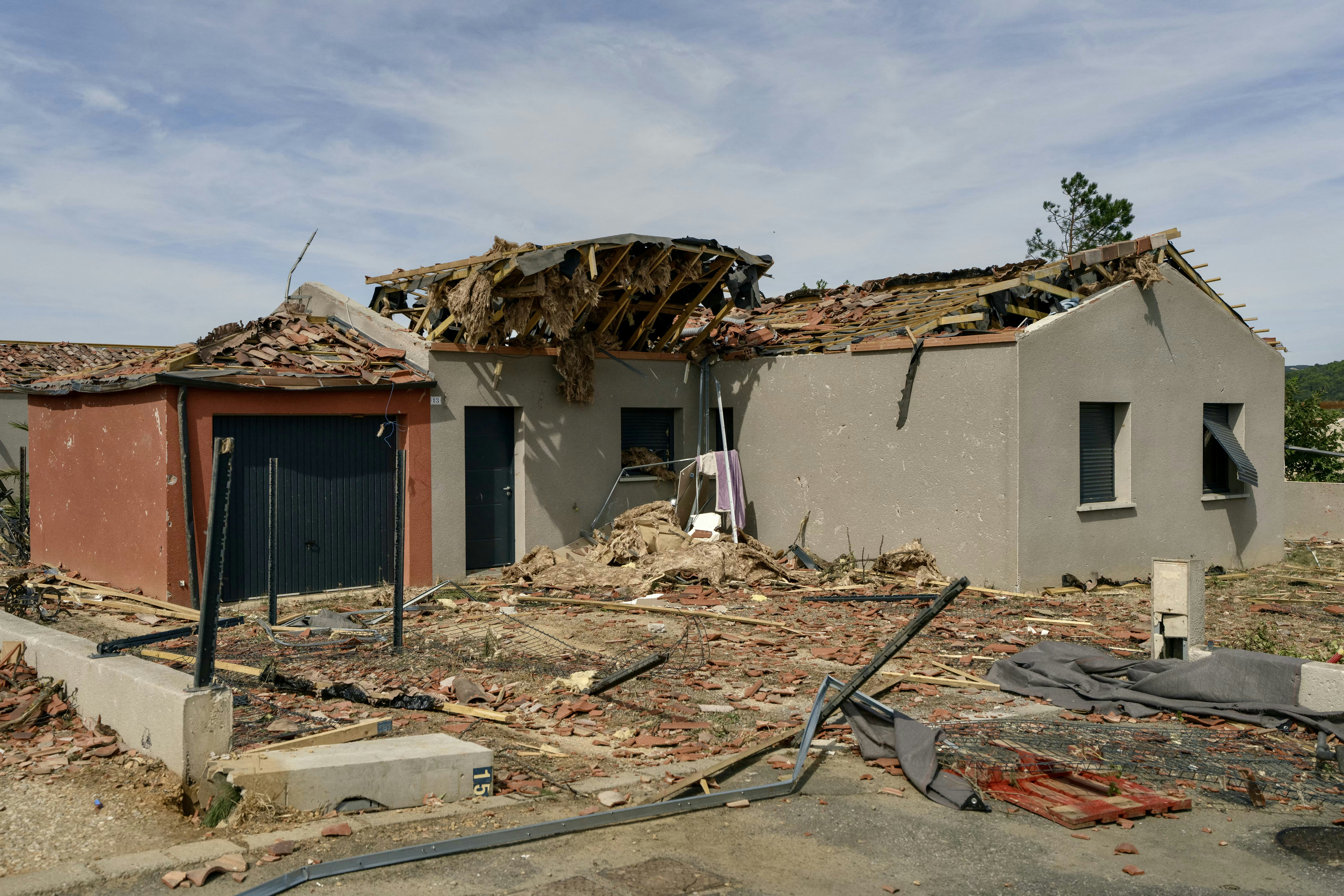 Duas casas com telhados destru&iacute;dos e escombros espalhados pelo ch&atilde;o, indicando danos por desastre natural. A casa da esquerda &eacute; marrom-avermelhada com porta de garagem preta, e a da direita &eacute; cinza-claro com janelas e portas escuras. O c&eacute;u est&aacute; parcialmente nublado