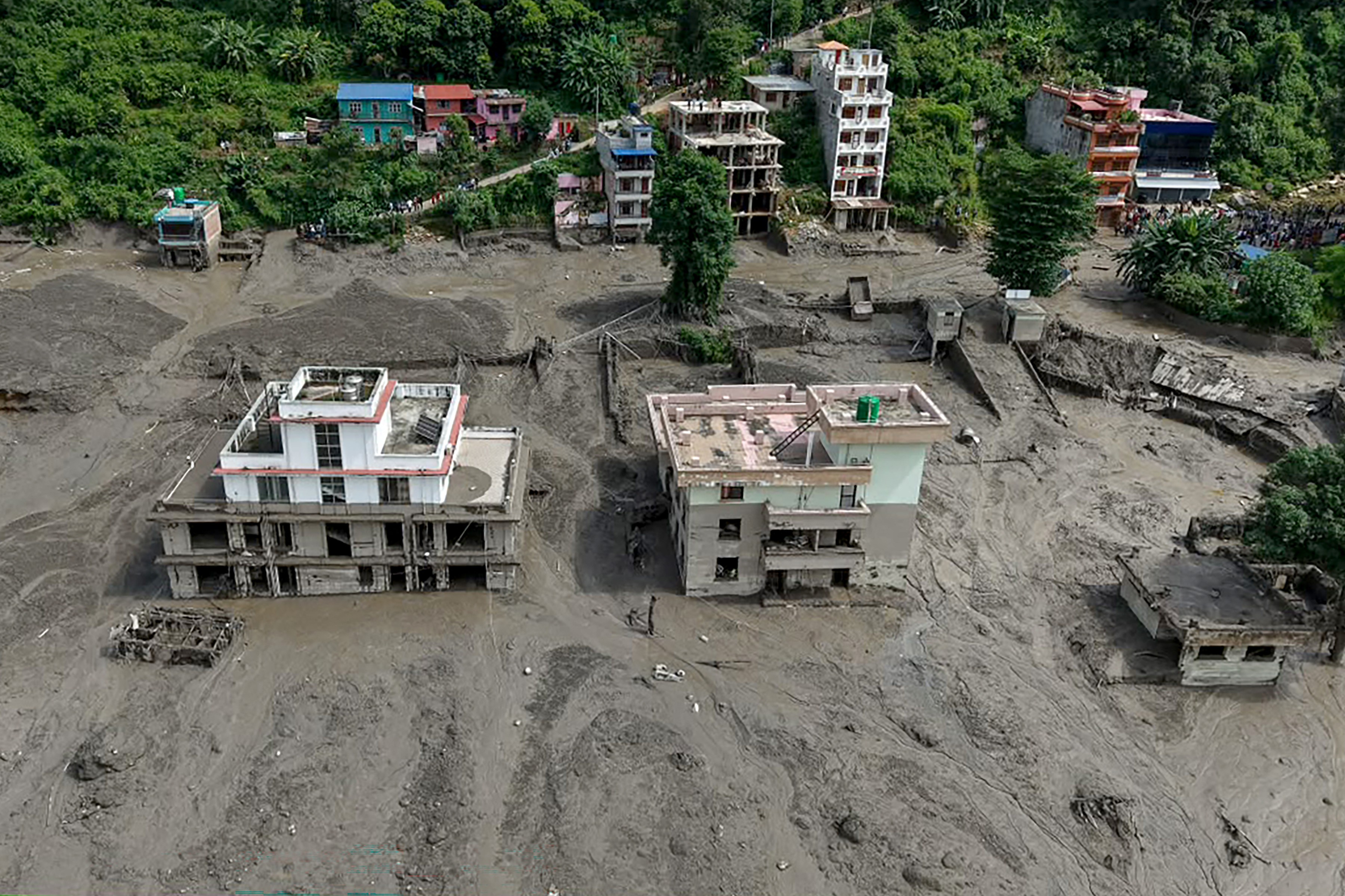 Vista aérea de uma cidade devastada por um deslizamento de terra, com casas parcialmente soterradas por lama cinza. Algumas construções estão intactas, outras desabadas ou com estruturas expostas. Ao fundo, casas coloridas e vegetação densa em uma encosta.