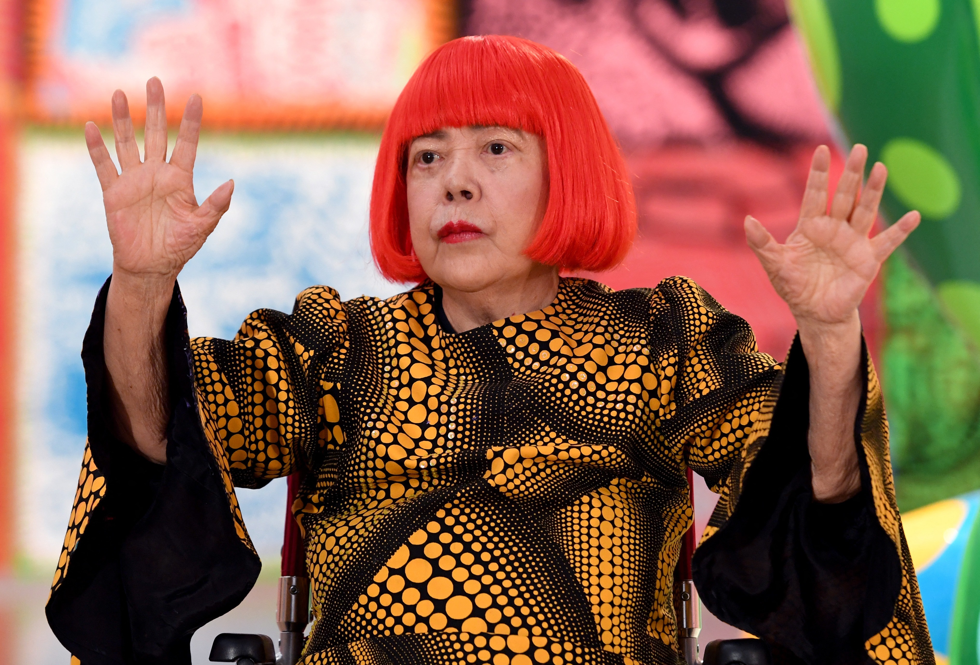 Artista Yayoi Kusama, com cabelo chanel vermelho vibrante e batom, veste blusa preta com bolinhas amarelas de tamanhos variados, levantando as mãos abertas em um gesto de saudação ou surpresa. Ela está sentada, com fundo desfocado em tons de rosa e verde