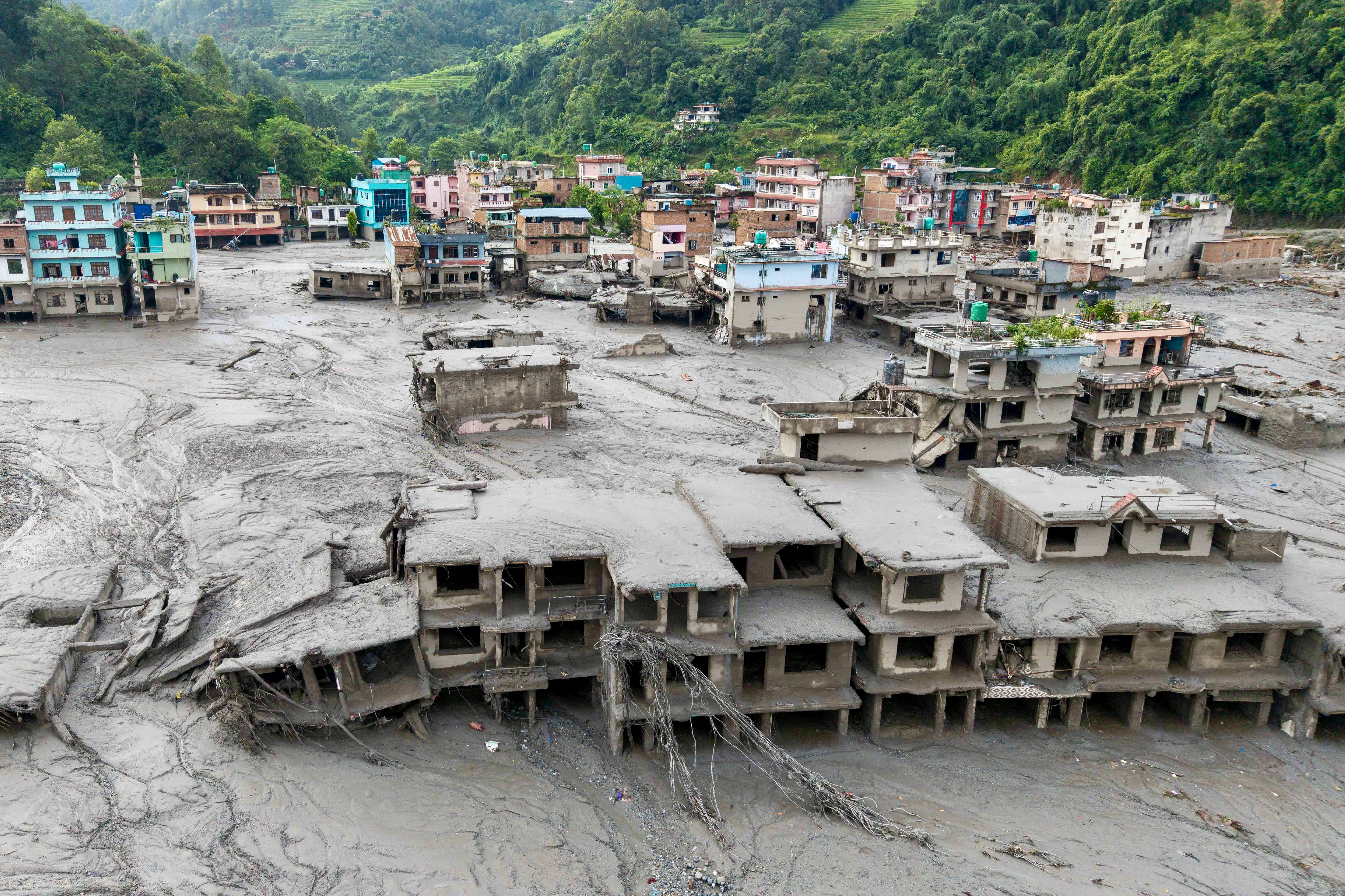 Vista aérea de uma cidade devastada por um deslizamento de terra, com edifícios parcialmente submersos em lama cinzenta. Algumas construções coloridas permanecem em pé ao fundo, enquanto as mais próximas estão destruídas e cobertas de detritos. Montanhas verdes e exuberantes formam o cenário