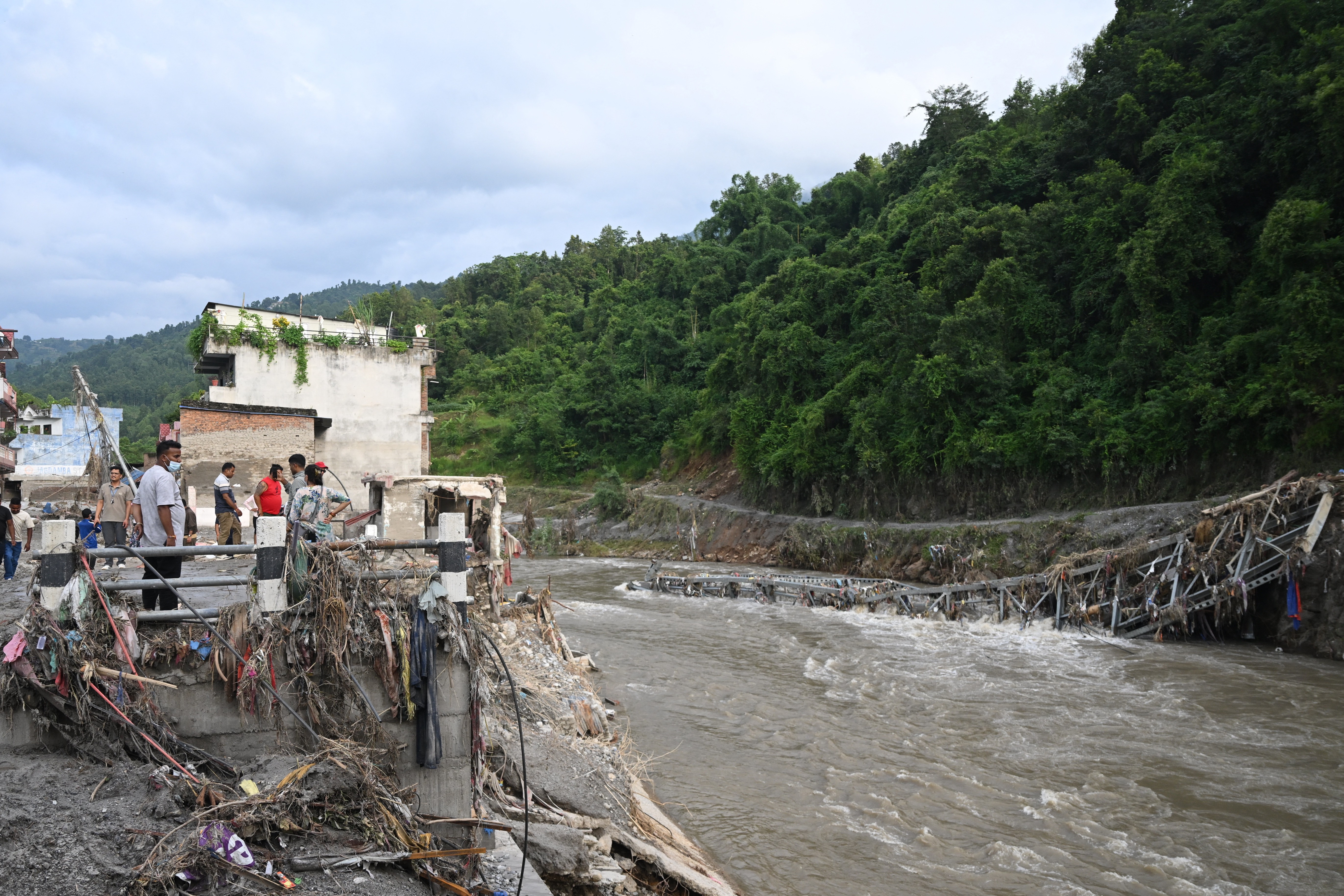 Pessoas observam os escombros de uma ponte destru&iacute;da por uma enchente, com detritos e lixo acumulados na margem do rio. Ao fundo, casas danificadas e uma montanha coberta por vegeta&ccedil;&atilde;o densa sob um c&eacute;u nublado
