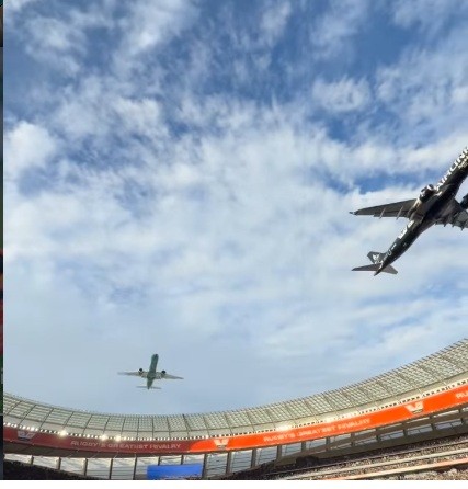 Dois aviões de passageiros voam sobre um estádio de futebol com arquibancadas vazias, sob um céu azul com nuvens esparsas