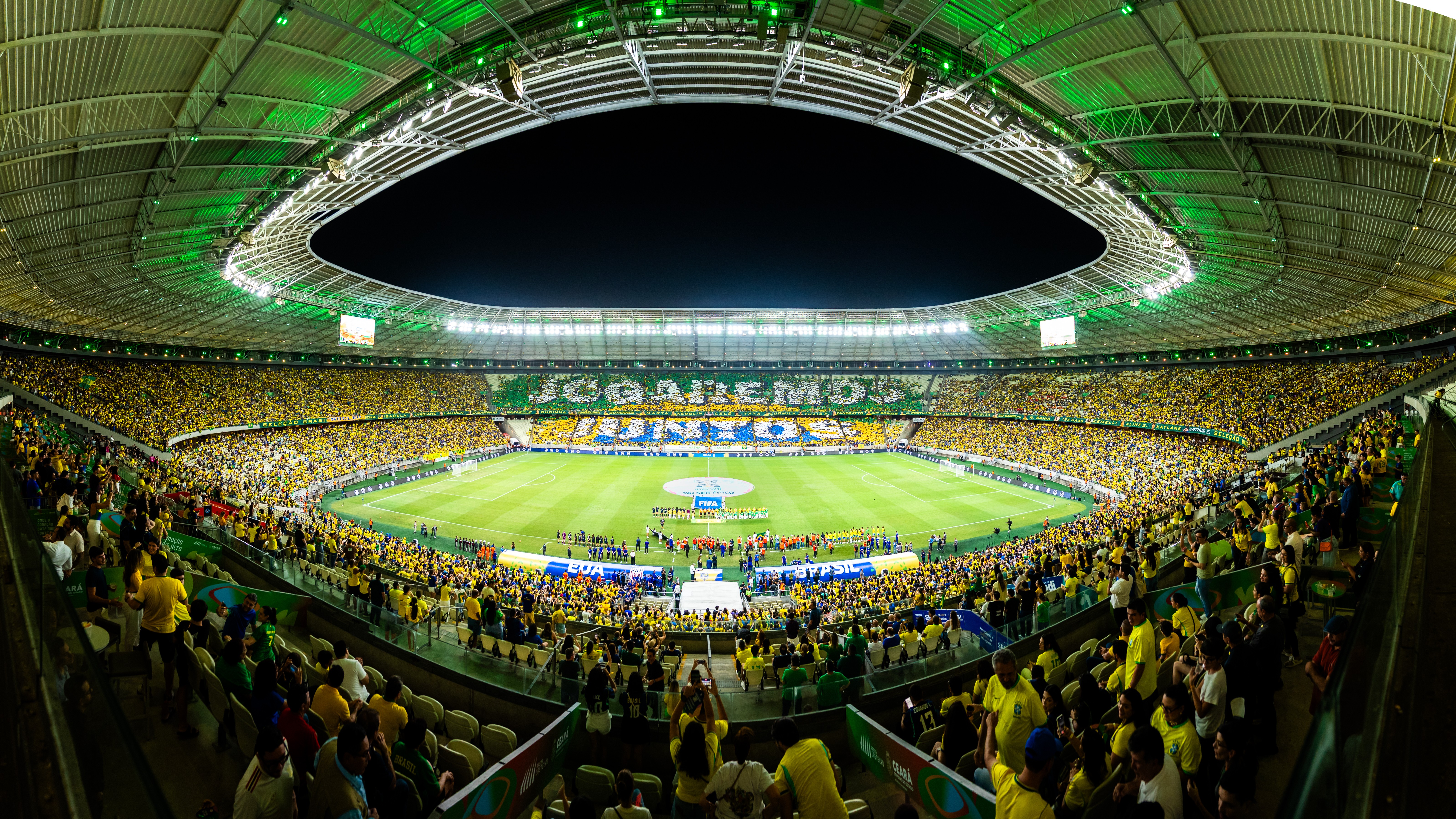 Est&aacute;dio de futebol lotado com torcedores vestindo amarelo e verde, iluminado por luzes verdes no teto. No campo, jogadores e equipe t&eacute;cnica se preparam para a partida. Nas arquibancadas, a frase JOGAREMOS JUNTOS &eacute; formada por torcedores com bandeiras e faixas