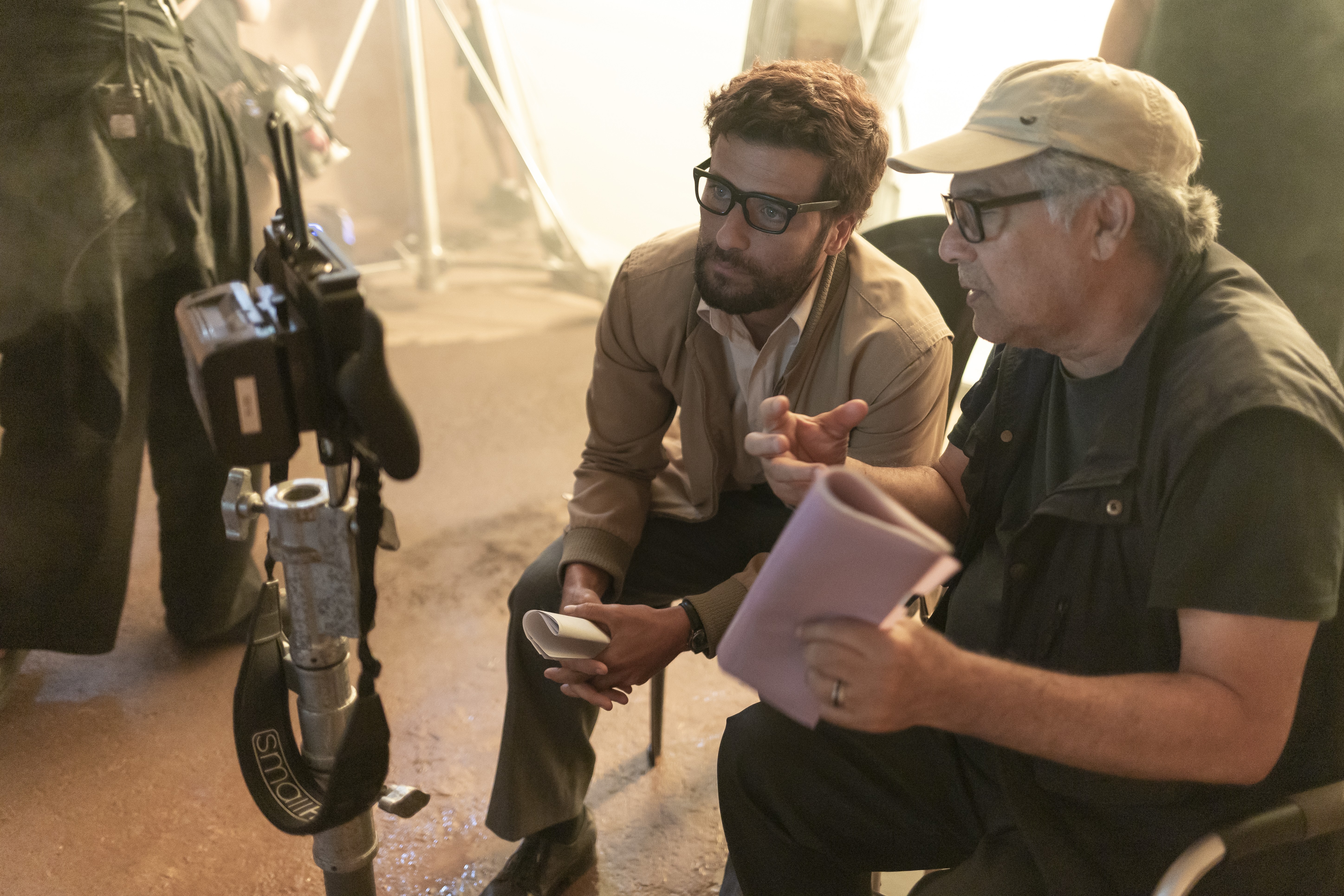 Dois homens sentados em um set de filmagem. À esquerda, um homem de óculos e jaqueta bege gesticula enquanto conversa com um homem de boné e colete escuro, que segura um roteiro roxo. Um equipamento de câmera está em primeiro plano à esquerda
