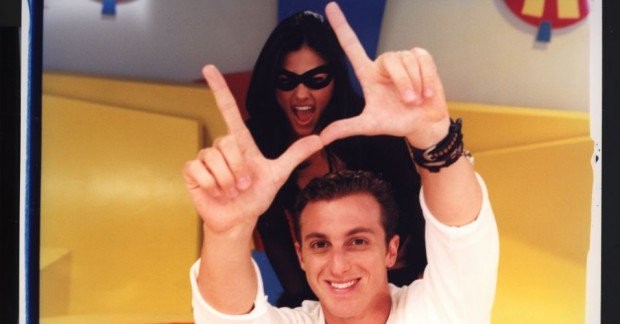 Luciano Huck sorrindo, com as mãos formando um L sobre a cabeça, e uma mulher mascarada de preto atrás dele, com a boca aberta em um grito, em um cenário amarelo