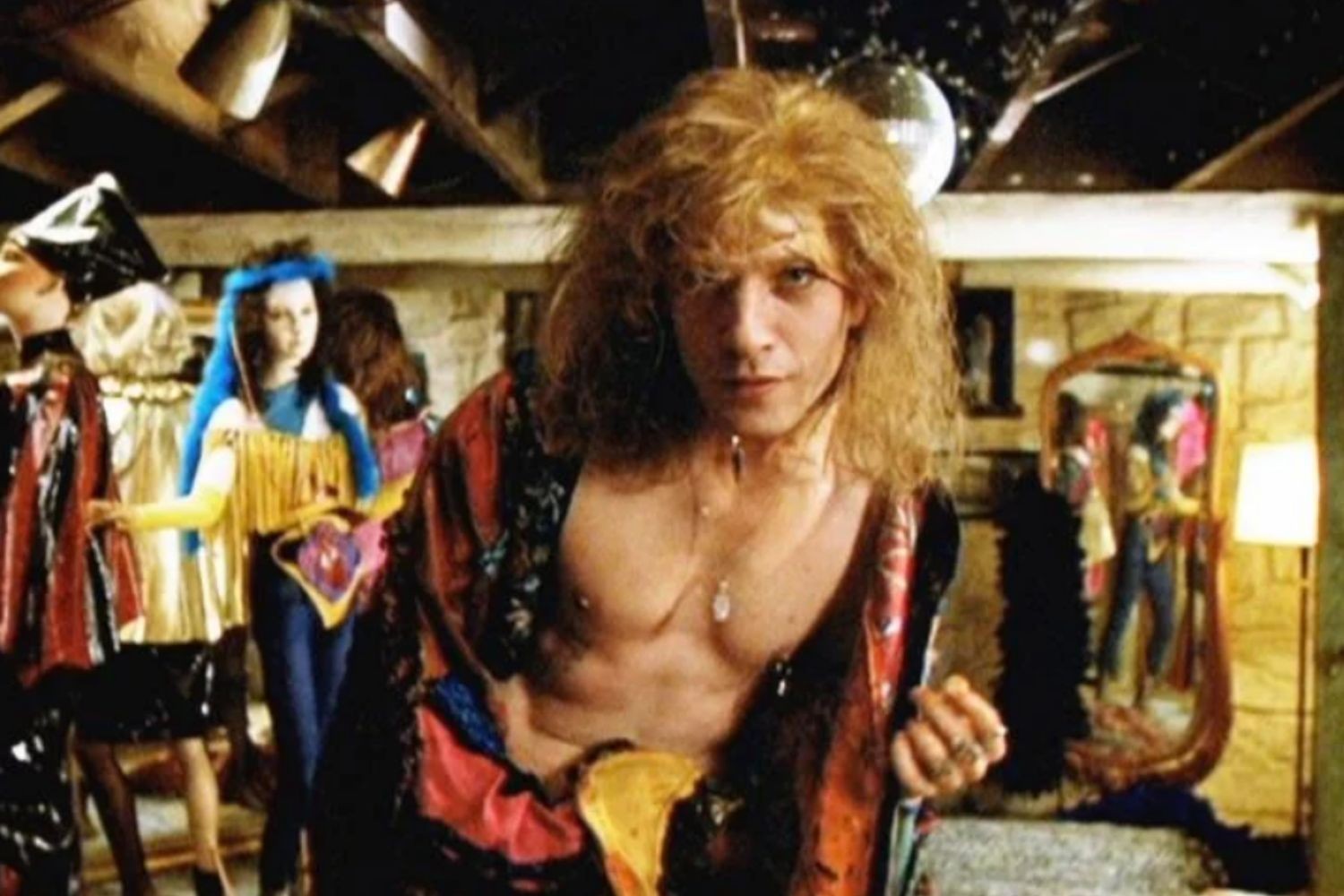 Homem de cabelos loiros e volumosos, sem camisa, usando um casaco colorido e colares, olha para frente. Ao fundo, manequins com roupas extravagantes e um espelho oval refletindo um ambiente com luzes e uma luminária de chão