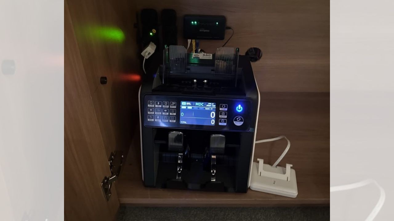 Máquina contadora de dinheiro preta e branca com tela azul ligada, exibindo MDC e zeros, dentro de um armário de madeira. Luzes verdes e vermelhas brilham no fundo, com cabos e um carregador branco ao lado