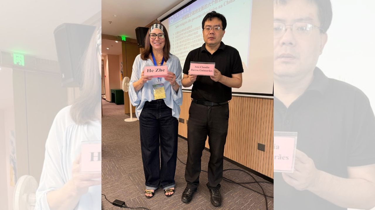 Mulher e homem sorrindo, de p&eacute;, segurando placas de identifica&ccedil;&atilde;o rosa com seus nomes, He Zhe e Jian Chuan, em um evento com projetor ao fundo
