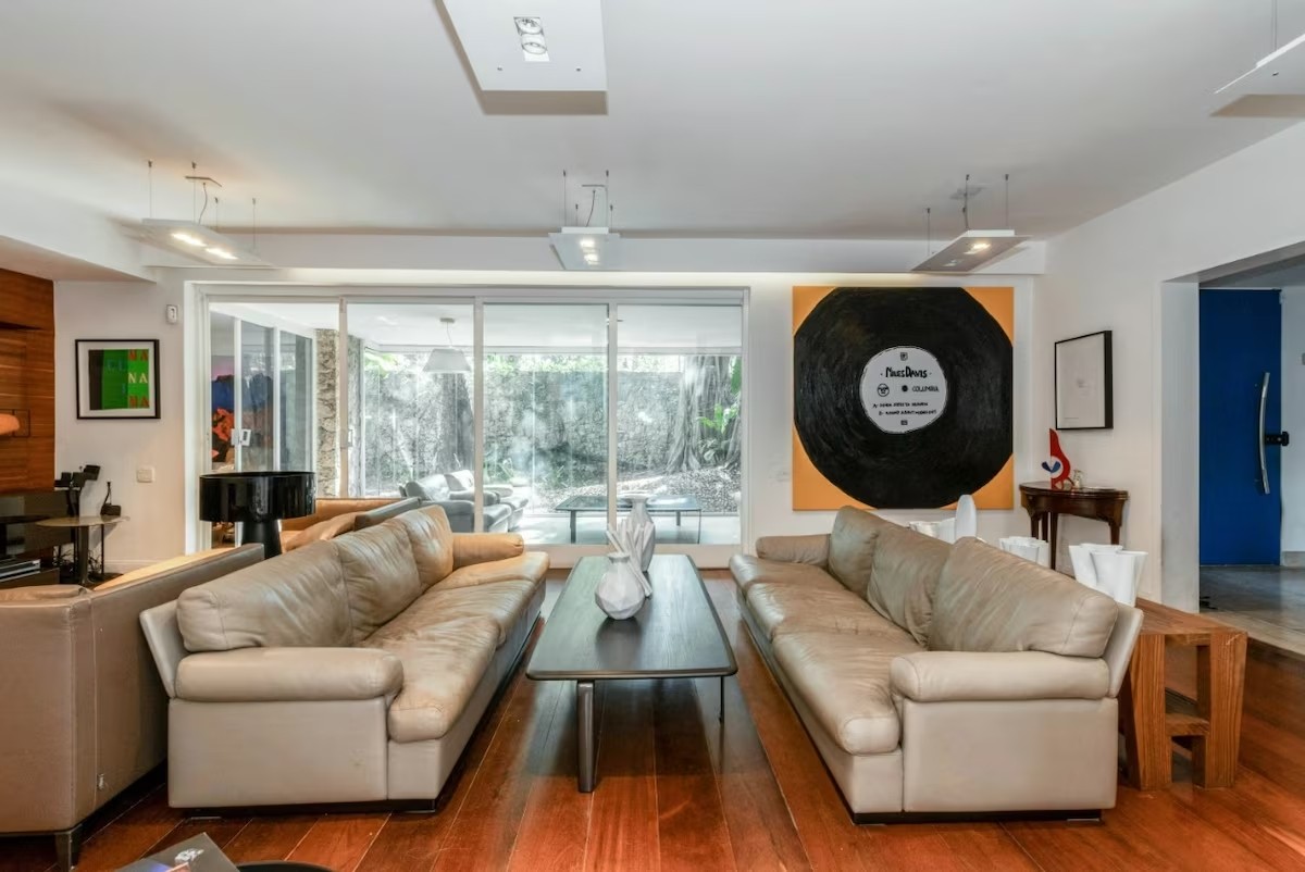 Sala de estar com dois sof&aacute;s de couro bege, um de frente para o outro, separados por uma mesa de centro escura com vasos brancos. Ao fundo, uma porta de vidro revela um jardim. Na parede, uma grande pintura de um disco de vinil preto sobre fundo laranja. &Agrave; direita, uma porta azul vibrante