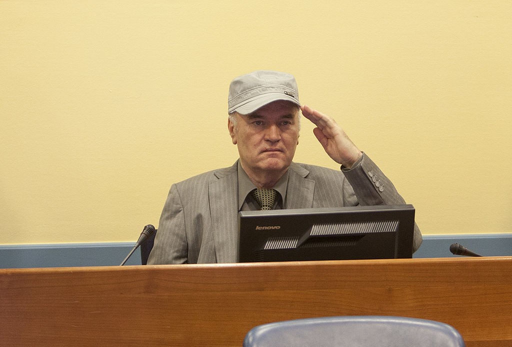 Homem de meia-idade, com boné cinza e terno risca de giz, faz saudação militar com a mão direita sobre a testa, sentado atrás de uma mesa de madeira com um monitor de computador à frente, em um ambiente com parede amarela