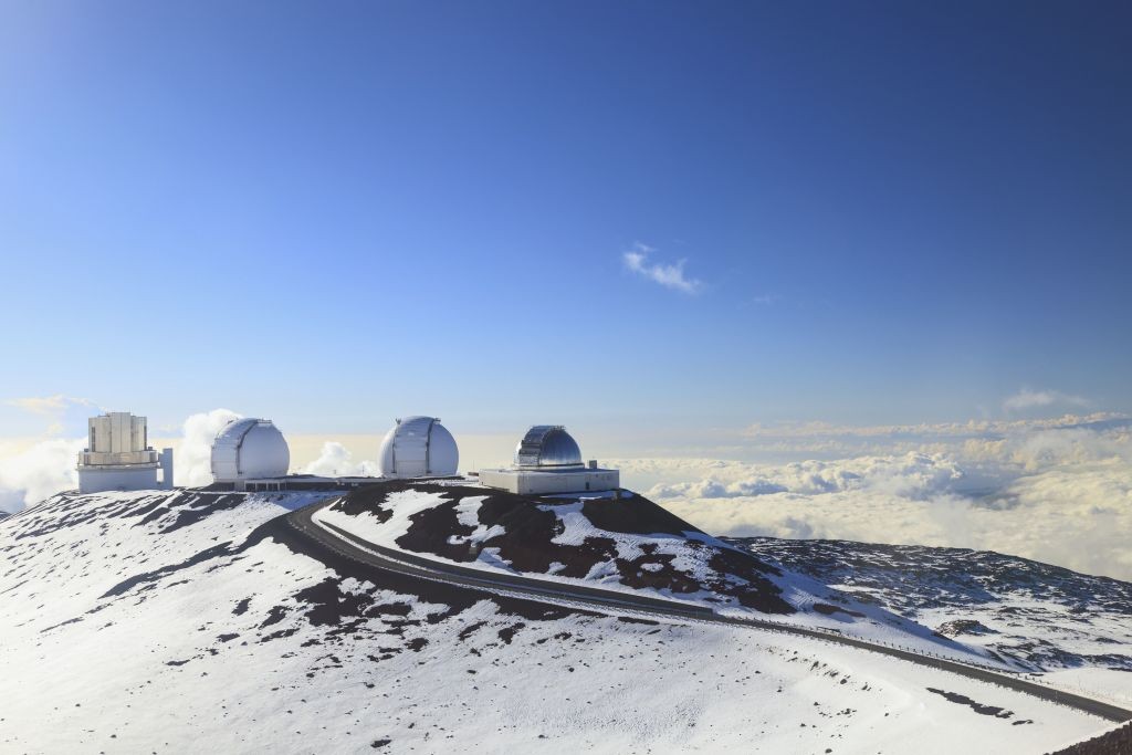 Quatro observatórios astronômicos brancos com cúpulas e um edifício retangular no topo de uma montanha coberta de neve, com uma estrada sinuosa e nuvens abaixo, sob um céu azul claro