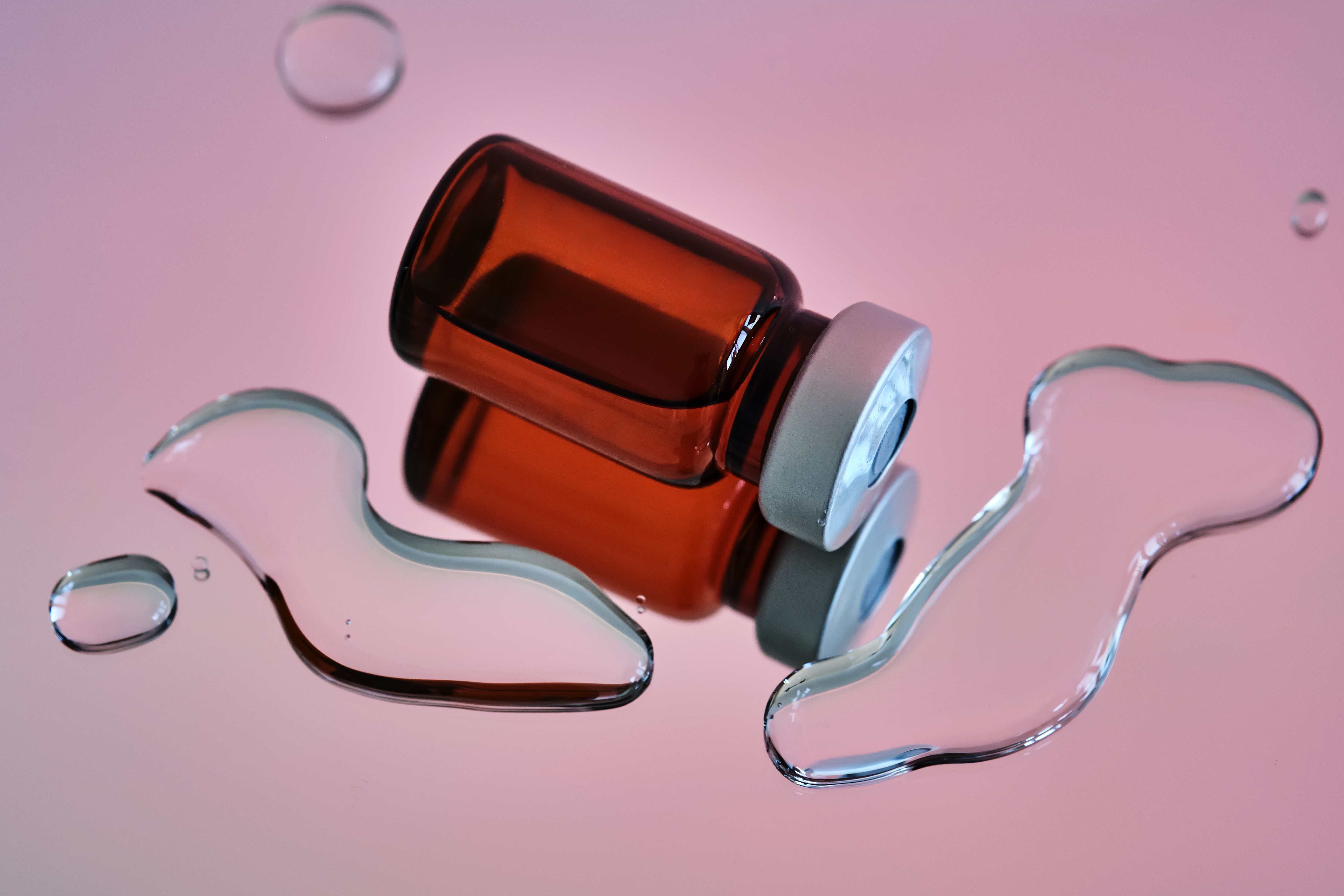 Frasco de vidro âmbar deitado sobre uma superfície rosa, refletindo-se. Gotas de água de tamanhos variados espalhadas ao redor, algumas maiores e com formatos irregulares, outras pequenas e redondas. O frasco tem uma tampa cinza e azul