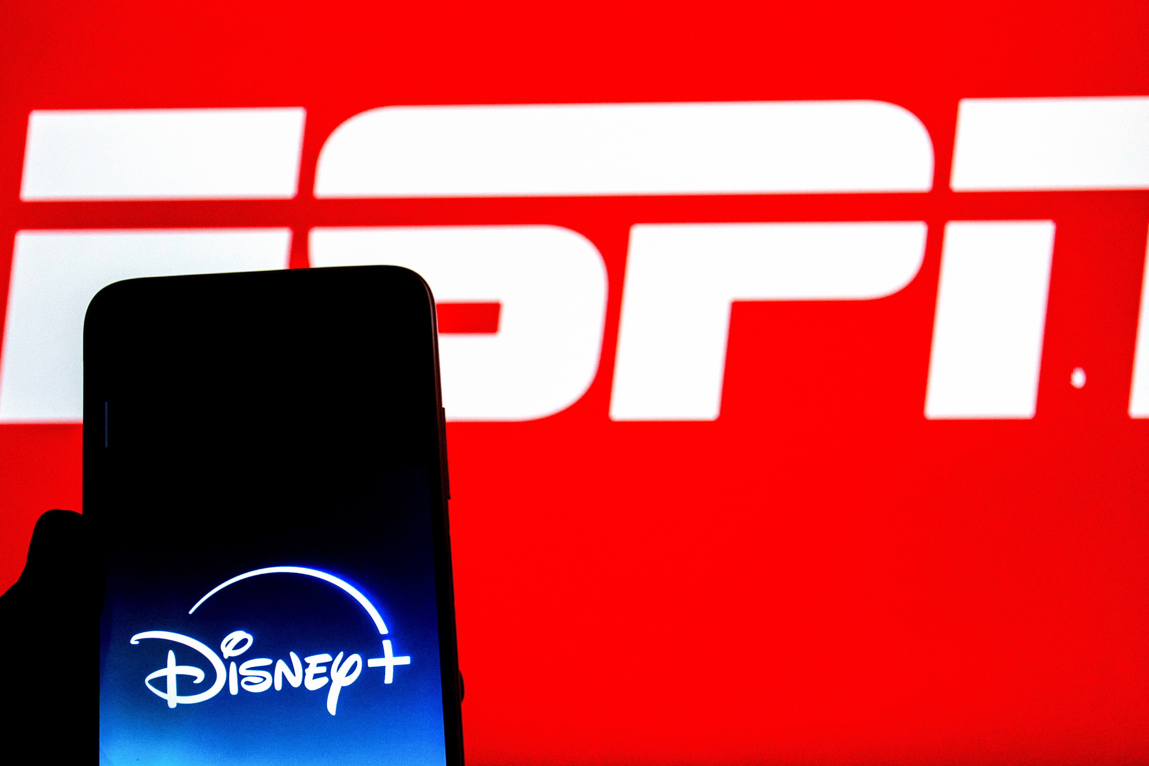 Um smartphone preto exibe o logotipo azul e branco do Disney+ em sua tela, com um fundo vermelho vibrante mostrando parte do logotipo branco da ESPN