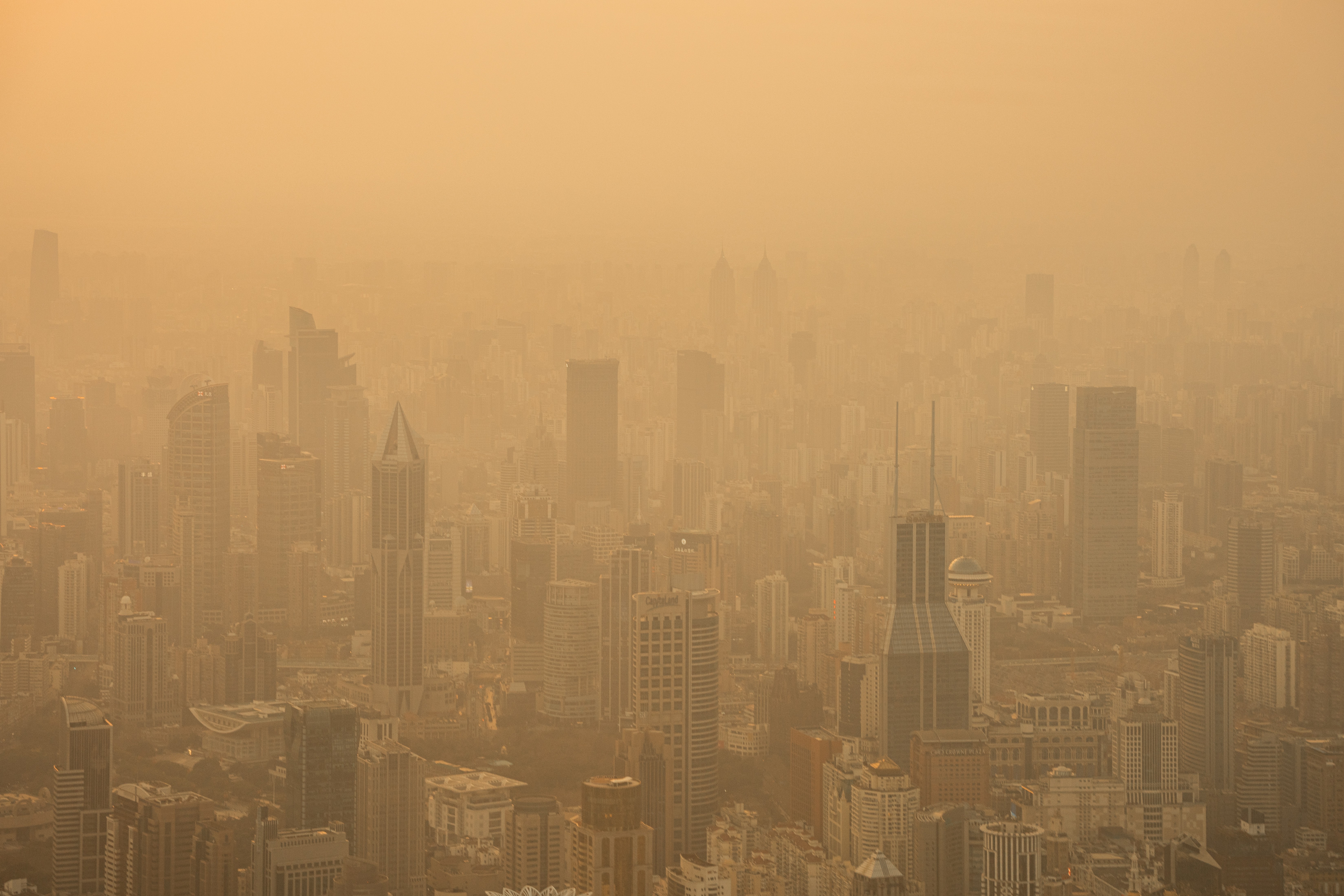 Vista aérea de uma cidade com muitos arranha-céus sob um céu e atmosfera densamente poluídos, com tons de laranja e marrom. A poluição obscurece a visibilidade, tornando os edifícios distantes quase imperceptíveis.