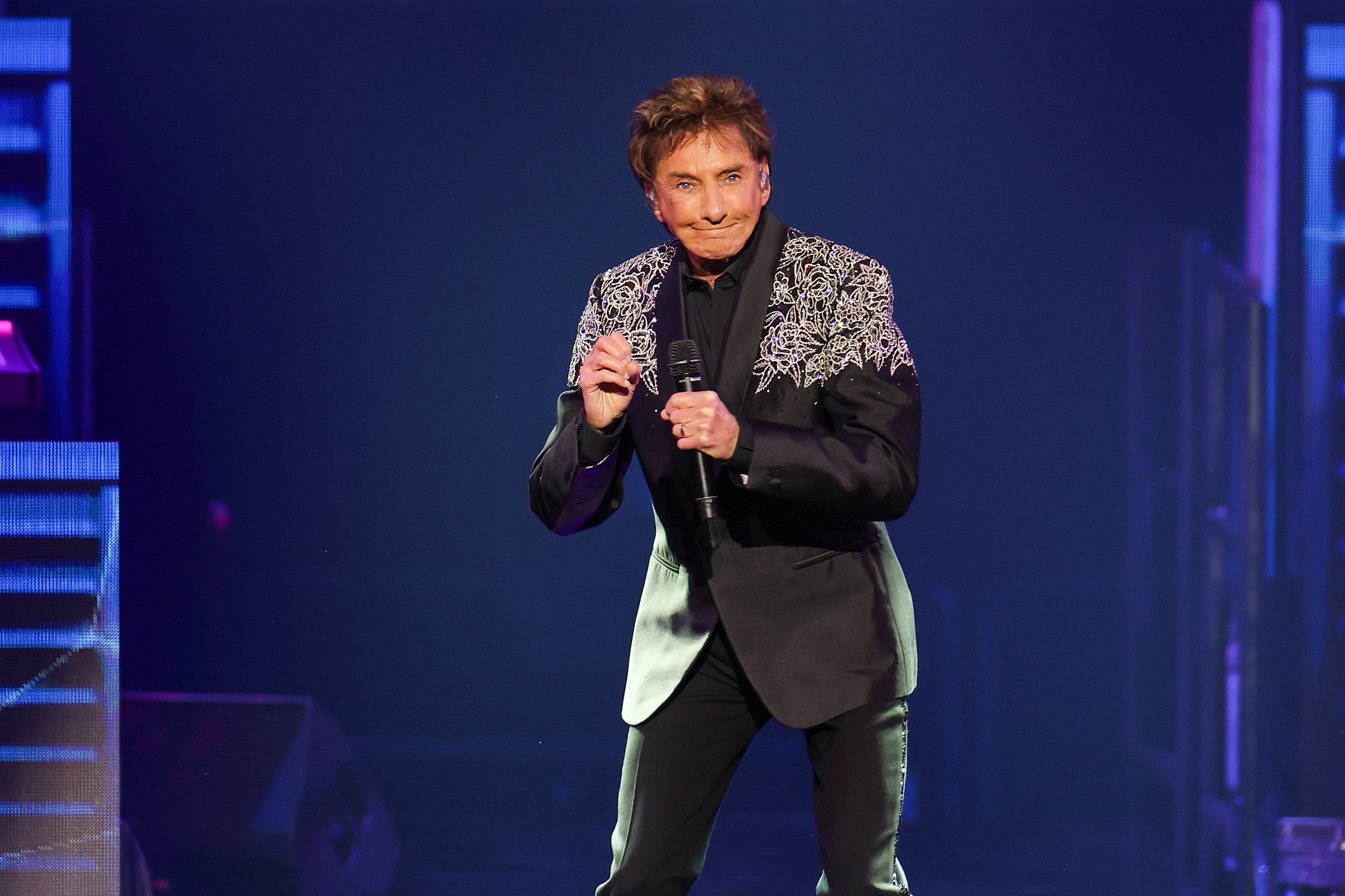 Cantor Barry Manilow, de cabelo castanho e terno preto com bordados prateados nos ombros, segura um microfone e gesticula no palco iluminado por luzes azuis
