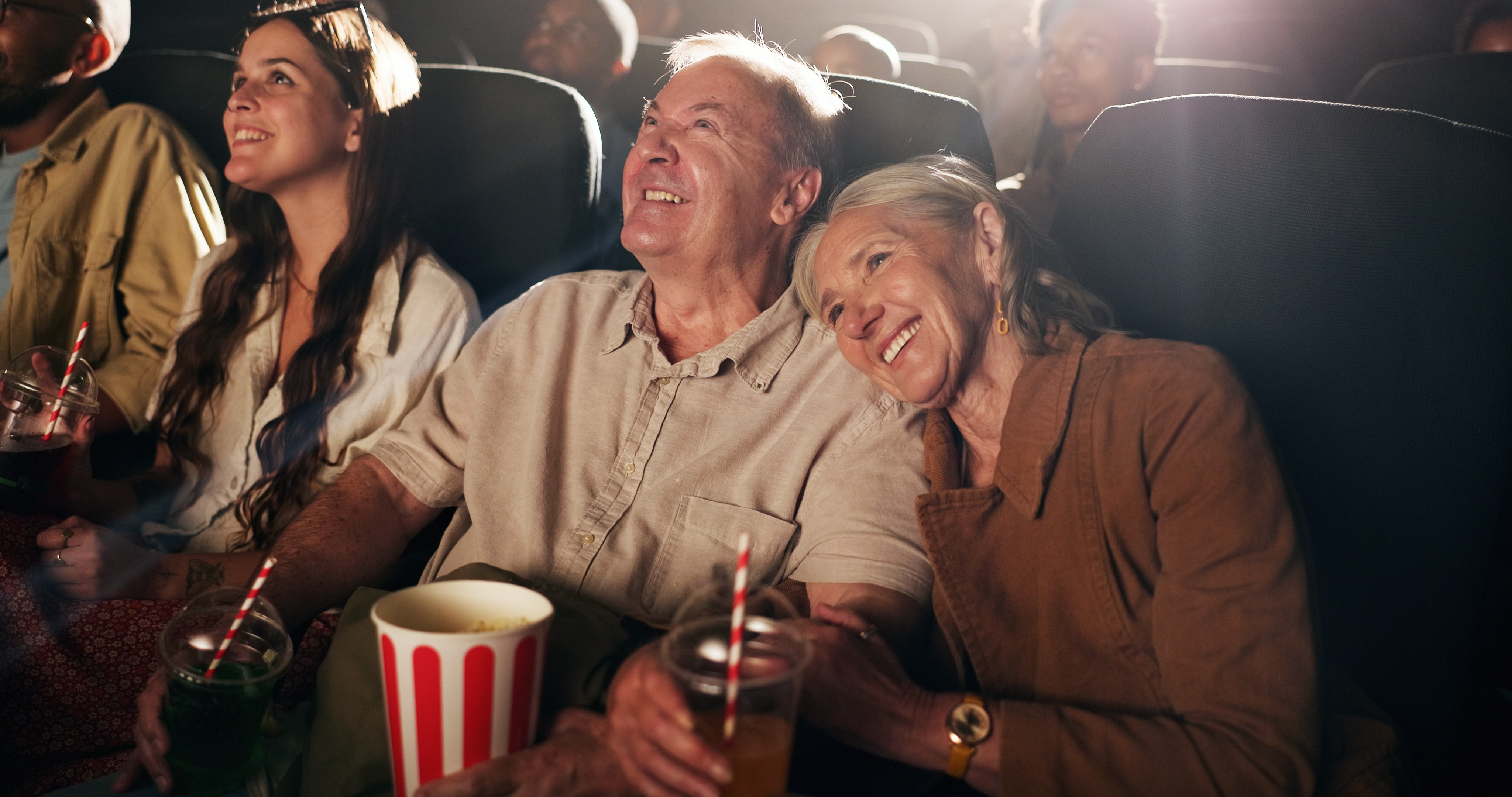 Casal idoso sorrindo e abraçado em um cinema escuro, com uma mulher mais jovem ao lado esquerdo também sorrindo. Eles seguram pipoca e refrigerantes, com a luz da tela refletindo em seus rostos felizes