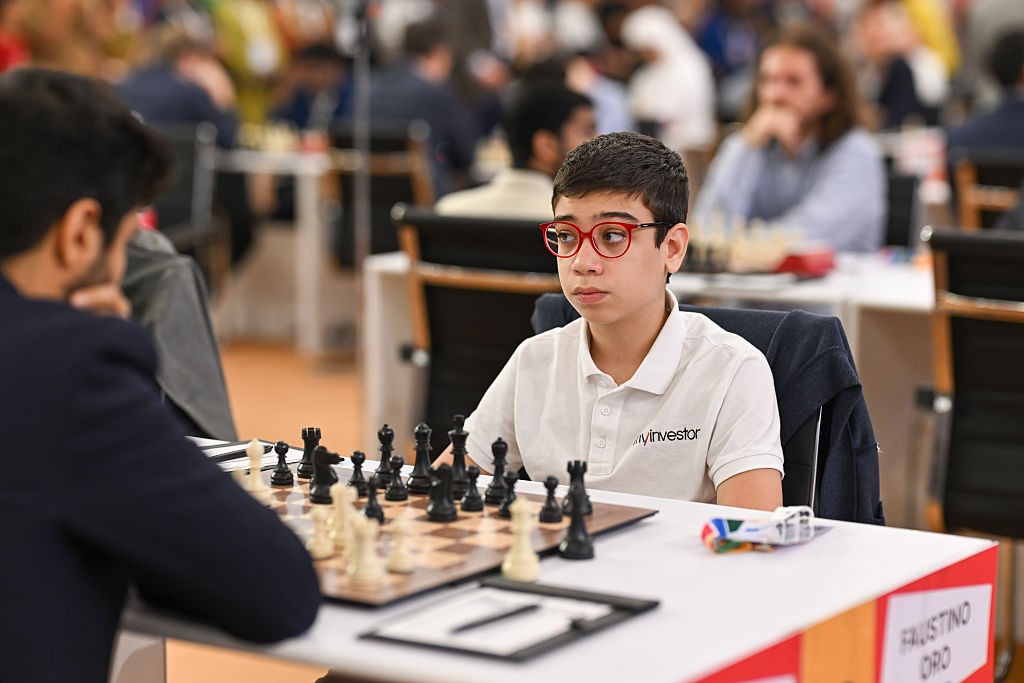 Jovem enxadrista de óculos vermelhos e camisa polo branca com logo Winvestor sentado à mesa, concentrado no tabuleiro de xadrez. Outro jogador, de costas, está à sua frente.