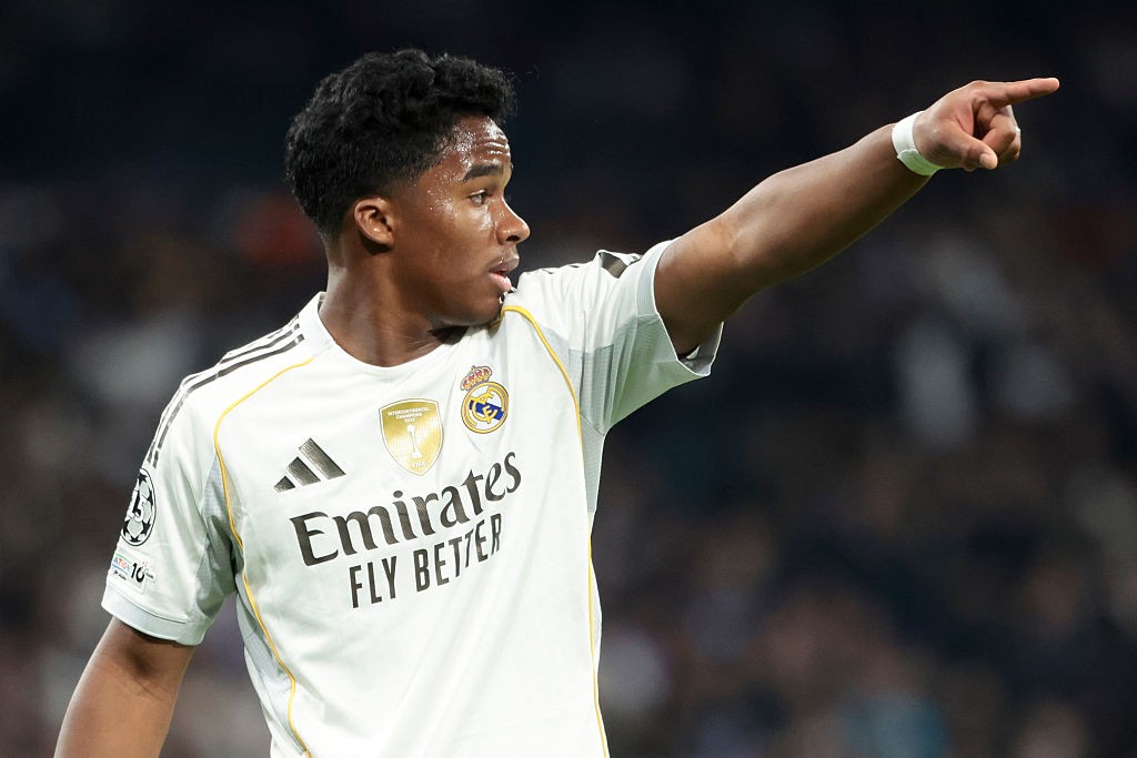Jovem jogador de futebol de pele escura, cabelo crespo, vestindo camisa branca do Real Madrid com detalhes dourados, apontando para a direita com o braço estendido e uma pulseira branca no pulso, em um estádio escuro