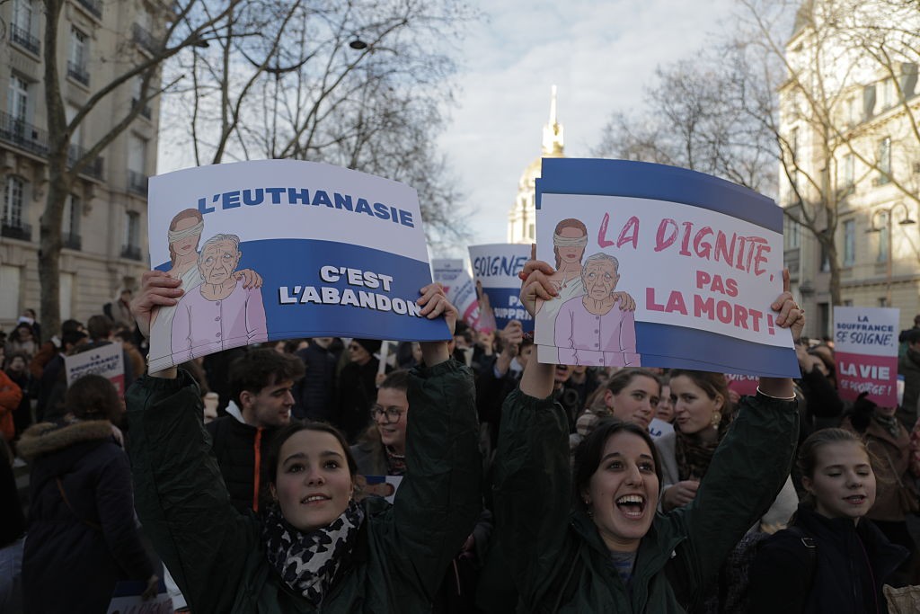 Duas mulheres jovens seguram cartazes contra a eutanásia em uma manifestação. O cartaz da esquerda diz Leuthanasie cest labandon (Eutanásia é abandono) e o da direita La dignité, pas la mort! (Dignidade, não a morte!). Ambos mostram uma pessoa vendada abraçando uma idosa triste.
