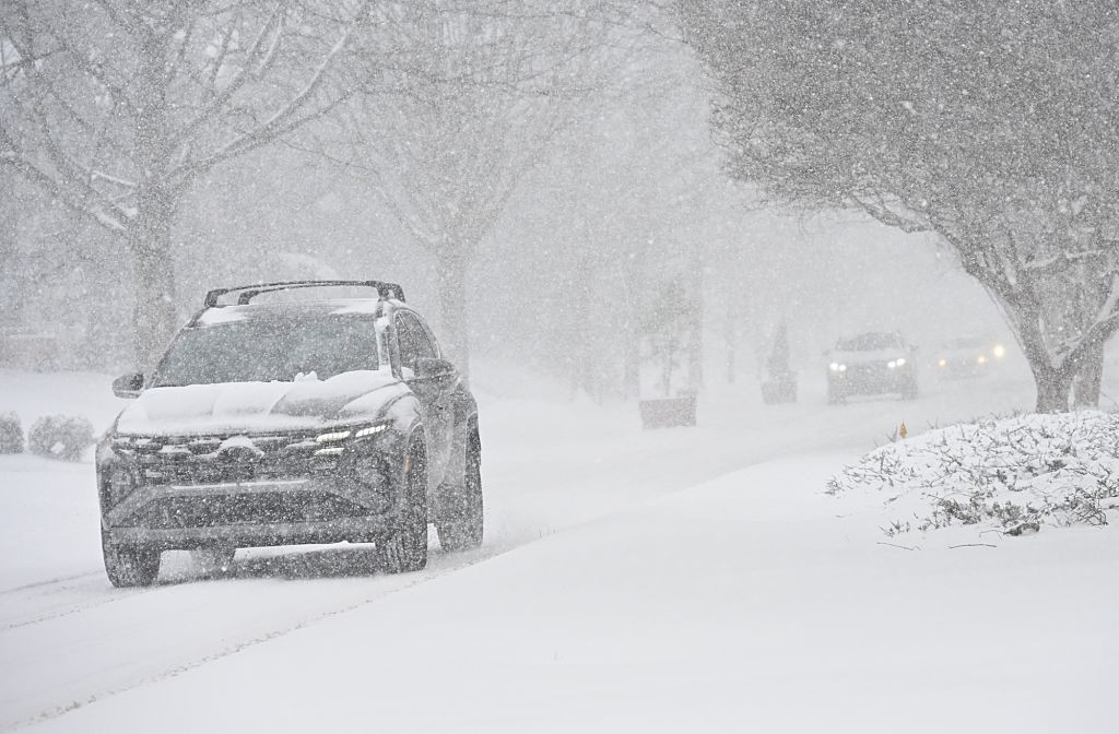 Carro escuro com bagageiro de teto trafegando em rua coberta por neve intensa, com árvores sem folhas e outros veículos ao fundo, em dia de nevasca