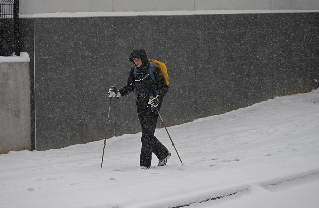 Pessoa agasalhada com casaco preto, capuz e mochila amarela, caminhando na neve com bastões de trekking, em dia de nevasca