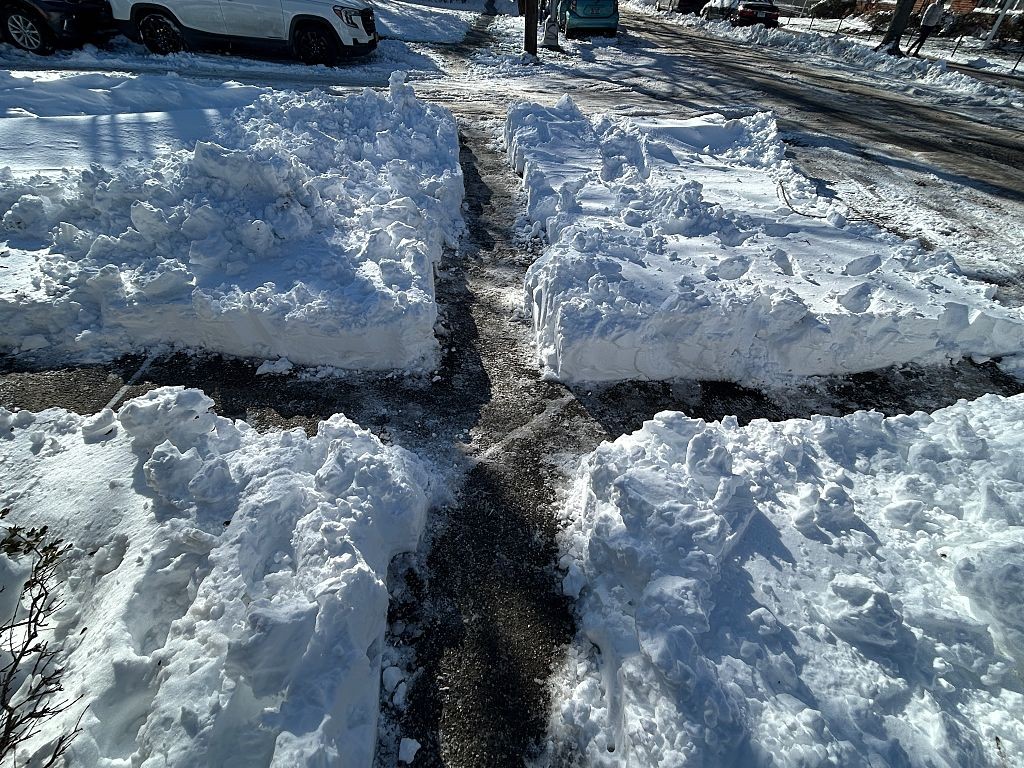 Caminho limpo de neve em formato de cruz, com montes altos de neve branca nas laterais, em uma rua com carros estacionados e pessoas ao fundo, sob sol forte
