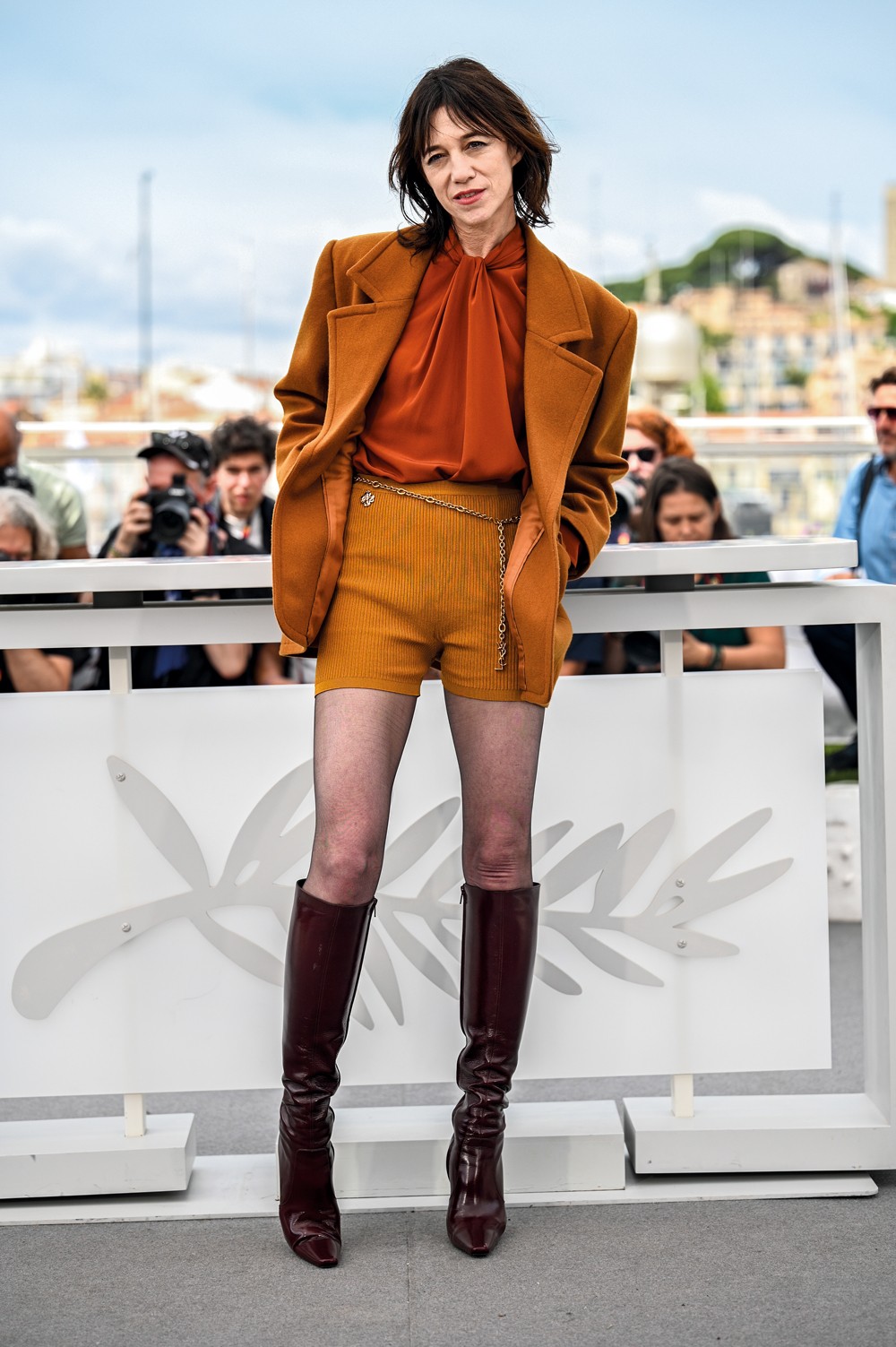 Charlotte Gainsbourg posa em evento ao ar livre, vestindo blazer marrom, blusa laranja, shorts mostarda com corrente dourada, meias-cal&ccedil;as pretas e botas de cano alto vinho. Ao fundo, pessoas e um cen&aacute;rio urbano desfocado