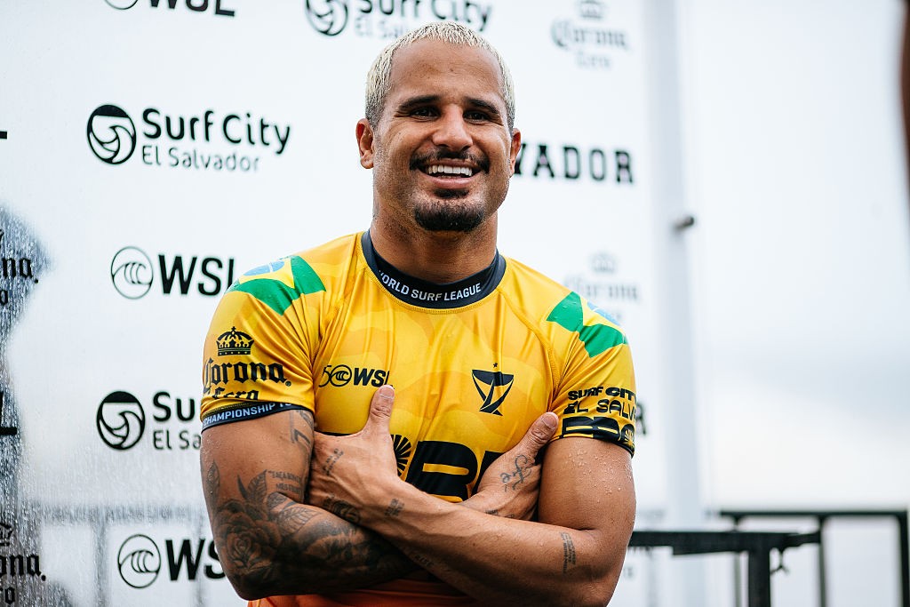 Homem sorridente com cabelo loiro e barba rala, vestindo camiseta amarela de surf com detalhes verdes e logotipos, braços cruzados, em frente a um painel branco com logotipos da Surf City El Salvador e WSL