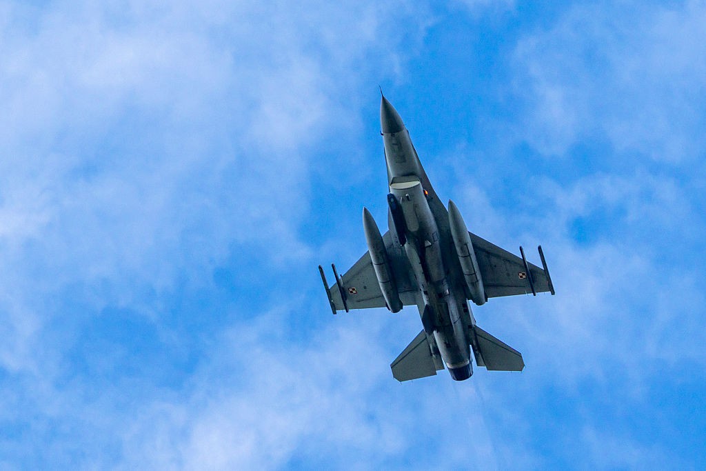 Caça militar cinza escuro visto de baixo, voando contra um céu azul com nuvens esparsas. O avião tem duas asas e um estabilizador vertical na cauda, com tanques de combustível ou mísseis sob as asas
