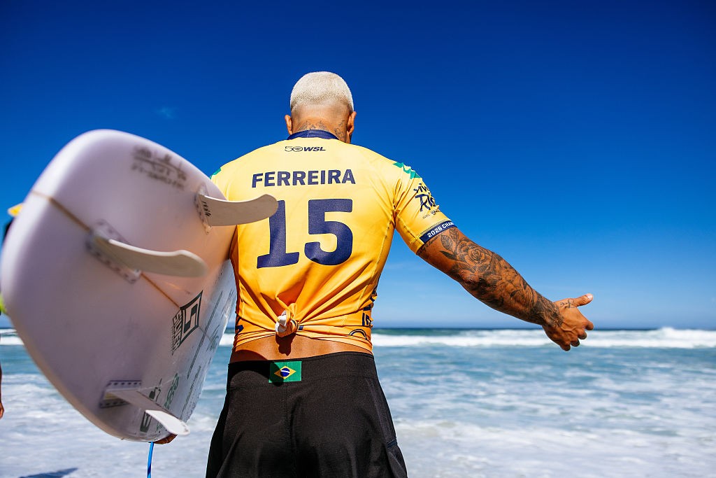 Um surfista de costas, com cabelo platinado, camiseta amarela FERREIRA 15 e bermuda preta com bandeira do Brasil, segura uma prancha branca e olha para o mar azul com ondas, sob céu claro