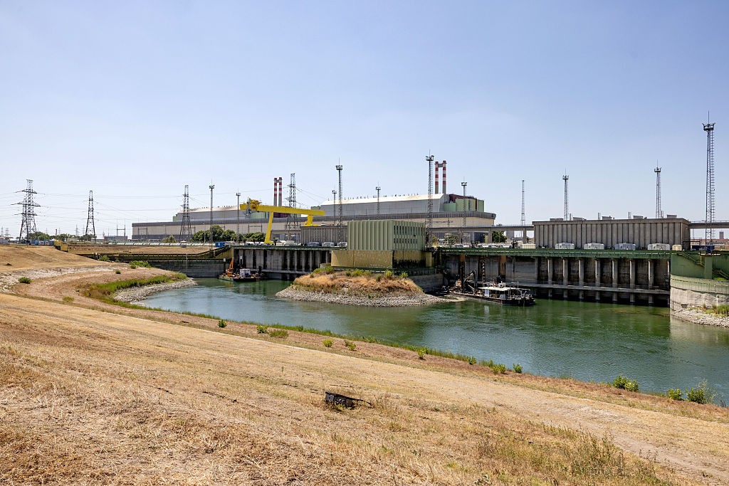 Usina hidrelétrica com grandes edifícios industriais cinzas e chaminés vermelhas e brancas ao fundo, com um rio verde-azulado em primeiro plano e margens de terra seca e grama amarelada. Um guindaste amarelo e torres de transmissão de energia completam a paisagem industrial sob um céu azul claro