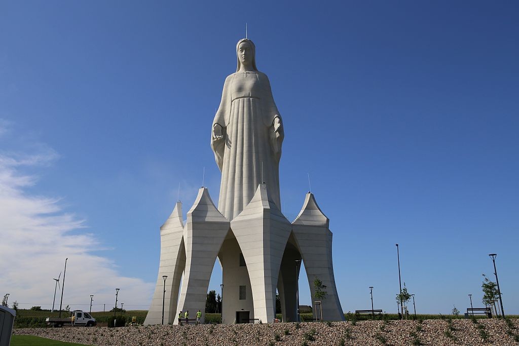 Estátua monumental de Nossa Senhora de Fátima, em concreto branco, com manto e braços abertos, sobre uma base arquitetônica moderna com arcos, sob céu azul claro. Ao redor, um terreno com pedras e vegetação rasteira, postes de luz e bancos. À esquerda, um caminhão branco e um mastro.