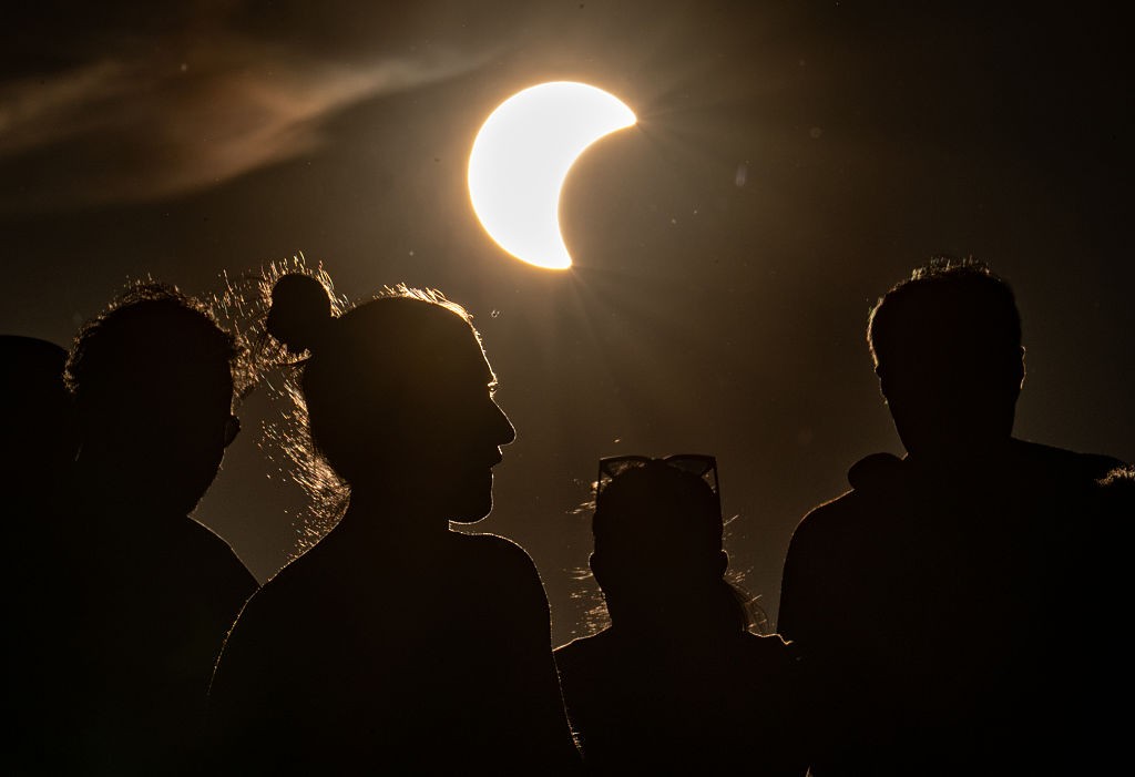 Silhuetas de pessoas observando um eclipse solar parcial, com o sol em formato de crescente brilhante no céu escuro, emitindo raios de luz