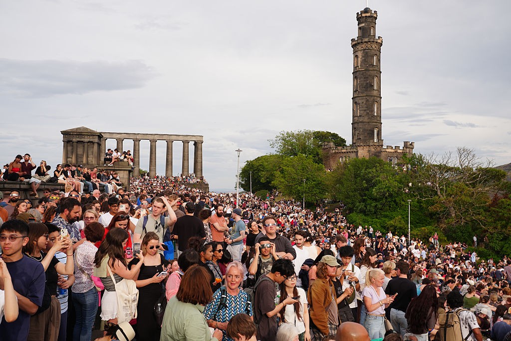 Multidão de pessoas diversas em um evento ao ar livre, com o Monumento Nacional da Escócia à esquerda e o Monumento a Nelson à direita, sob céu nublado