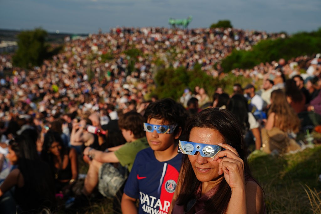 Mulher e menino usam óculos especiais para observar um eclipse solar, sentados em uma colina gramada com uma multidão ao fundo