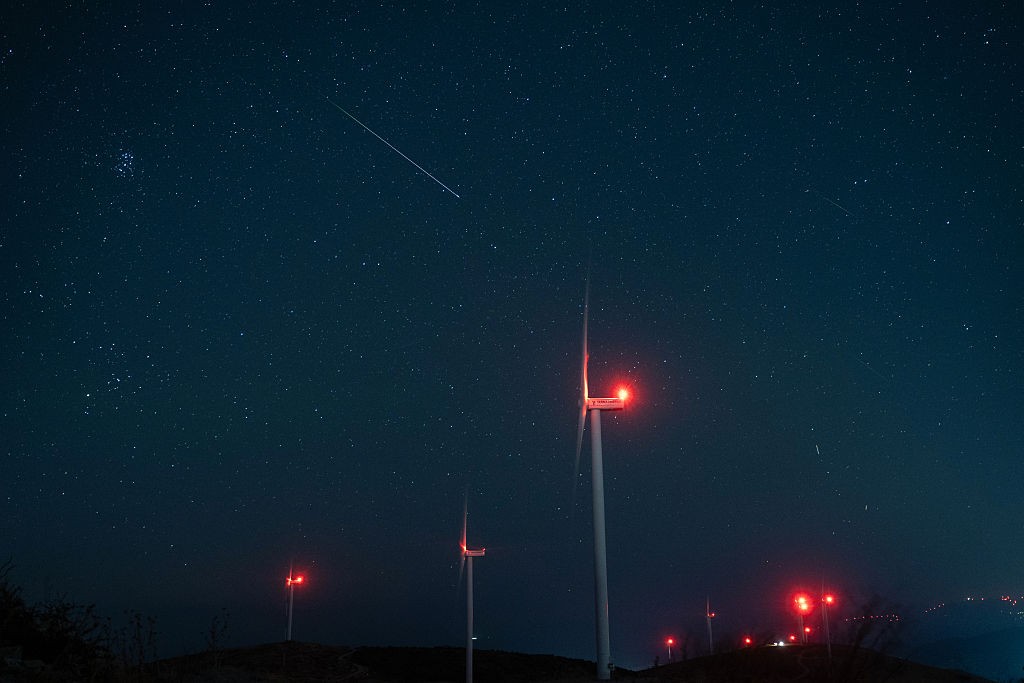Céu noturno estrelado com uma estrela cadente diagonal e vários aerogeradores com luzes vermelhas piscando no topo de uma colina escura