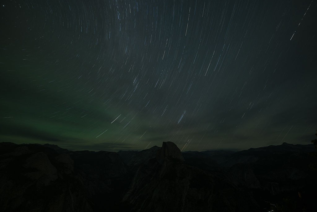 Céu noturno escuro com rastros de estrelas brancas e finas cruzando a imagem, e uma faixa verde de aurora boreal no horizonte. Abaixo, montanhas escuras e vales profundos, com um pico rochoso proeminente no centro-direita