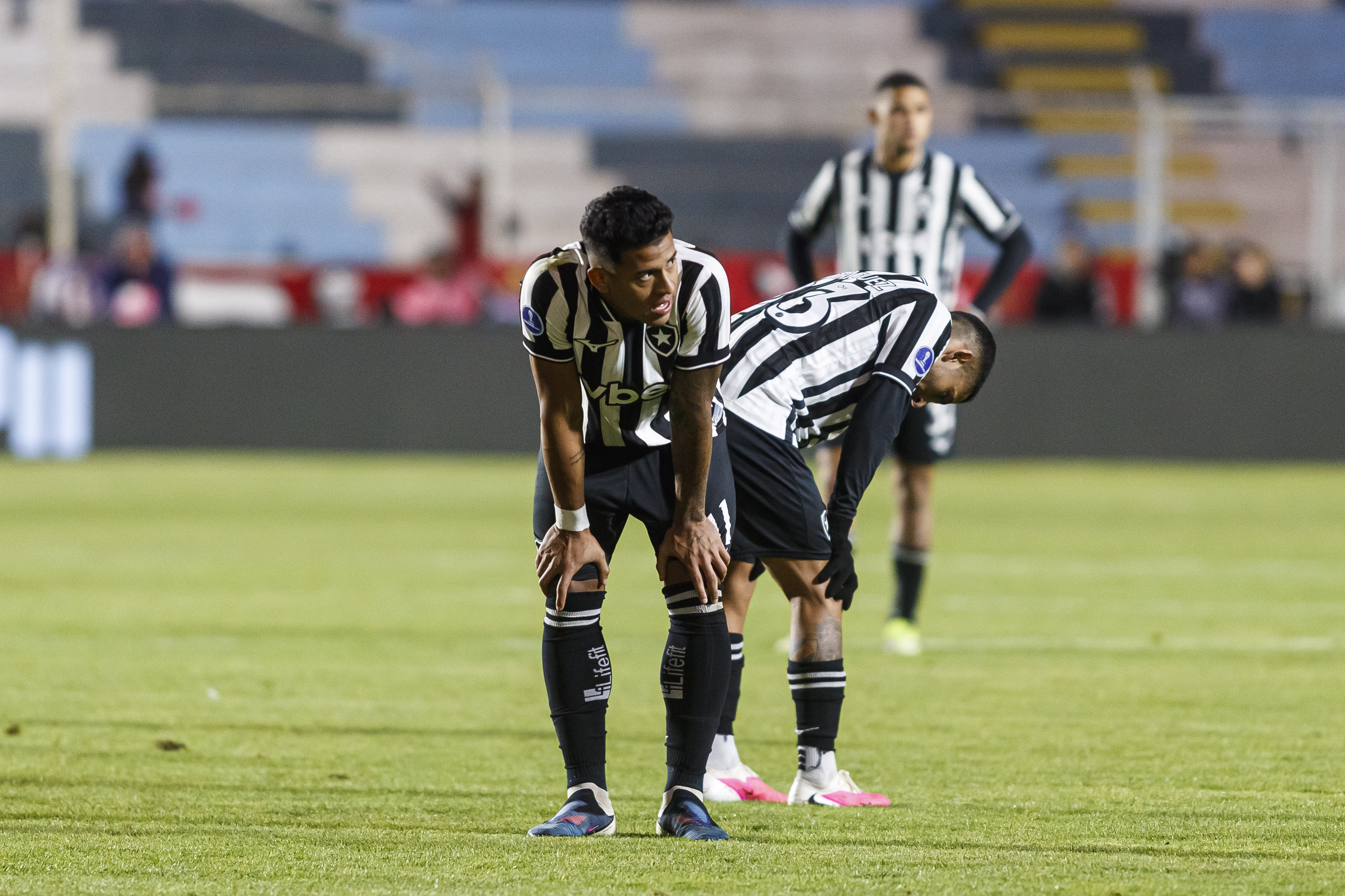 Três jogadores de futebol em campo, vestindo uniformes listrados em preto e branco. Dois estão curvados com as mãos nos joelhos, parecendo cansados, e um terceiro está mais ao fundo, em pé