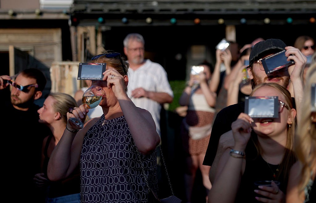 Pessoas observam um eclipse solar usando óculos especiais e filtros em celulares. Uma mulher no centro bebe vinho enquanto segura um filtro.
