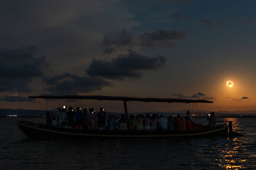 Um barco escuro, com um grupo de pessoas sentadas, navega em águas calmas ao entardecer. O céu, em tons de azul escuro e laranja, exibe um eclipse solar anular, com um anel de luz dourada ao redor da lua.