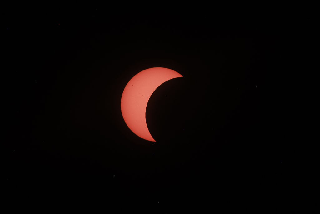 Eclipse solar parcial, com o Sol em formato de crescente laranja-avermelhado, visível contra o céu escuro pontilhado por pequenas estrelas