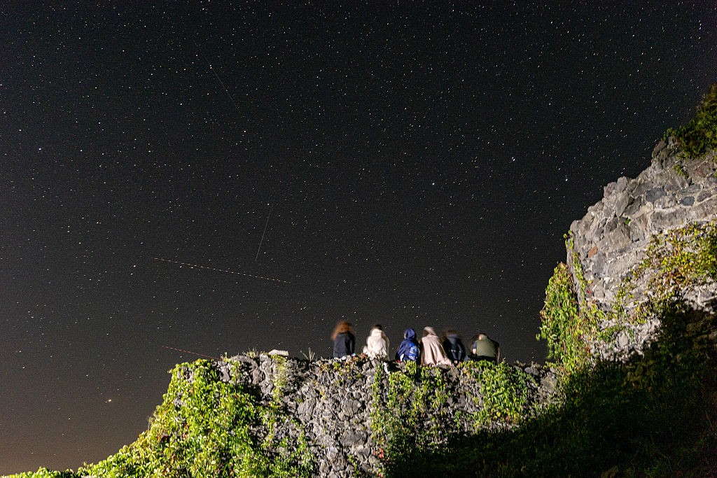 Seis pessoas sentadas de costas em um muro de pedra coberto por hera, observando um céu noturno escuro repleto de estrelas e rastros de luz
